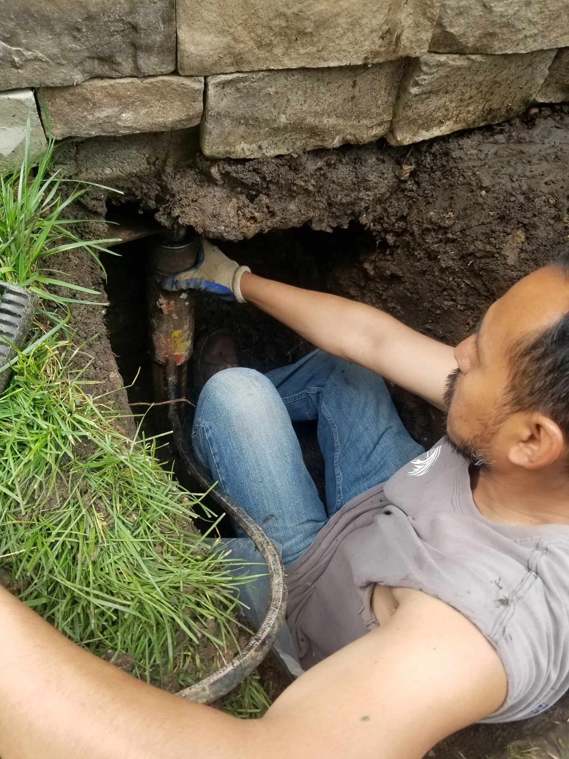 Man repairing underground pipe in a trench with soil and stone wall nearby, holding the pipe with gloves.