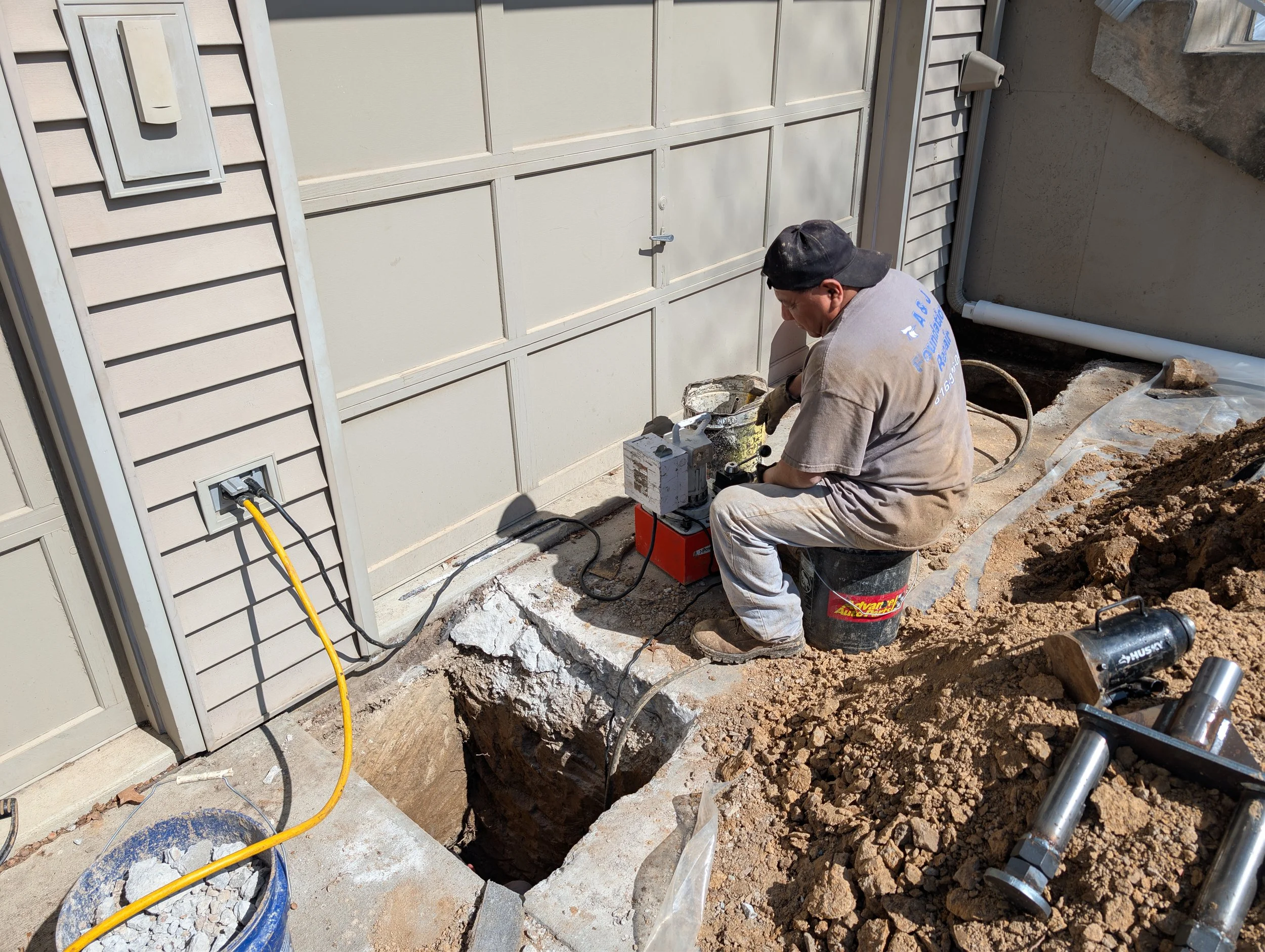 A construction worker operating a concrete breaking tool next to the exterior wall of a house, with dirt and rubble around.