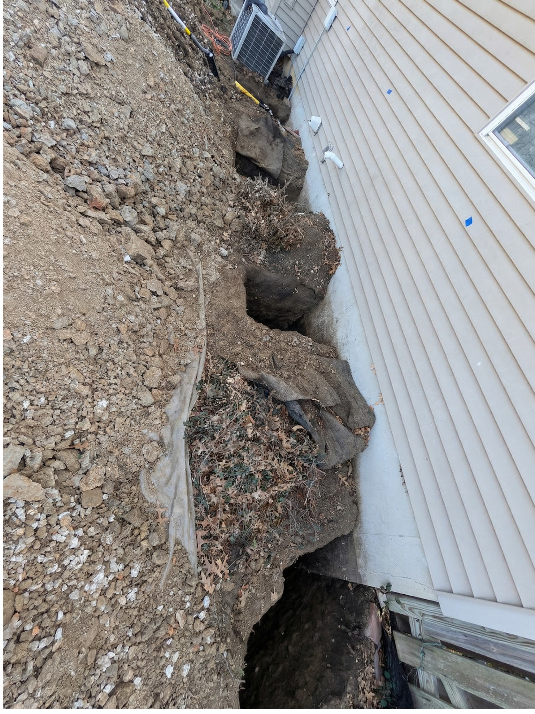 Excavation next to a house, showing dirt, rocks, and holes in the ground, with an air conditioning unit and house siding in the background.