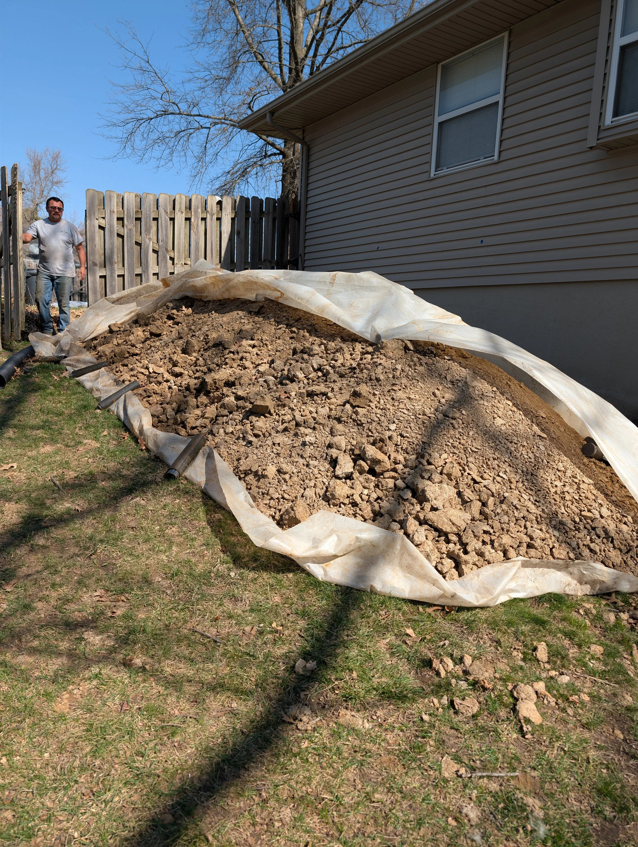A backyard with an uneven dirt excavation area lined with a plastic sheet, a man standing near a wooden fence, a house with beige siding, and leafless trees in the background.