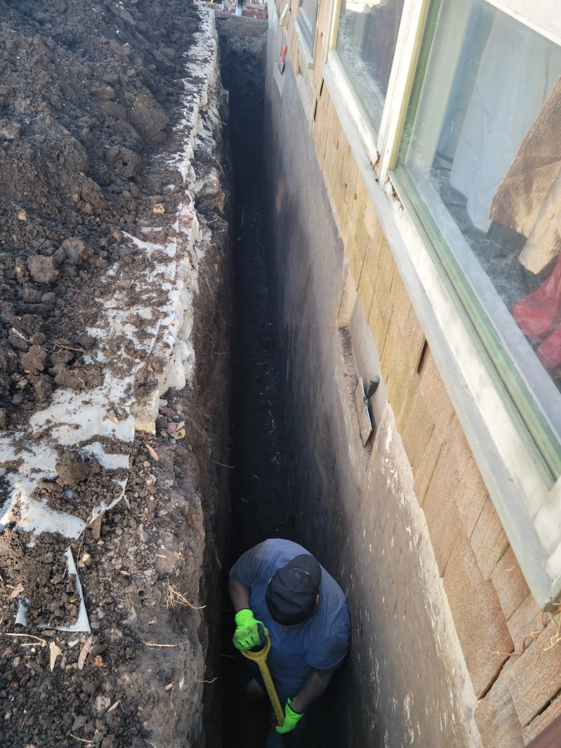 Worker digging a trench next to a house foundation, with a shovel, wearing gloves and a hat.