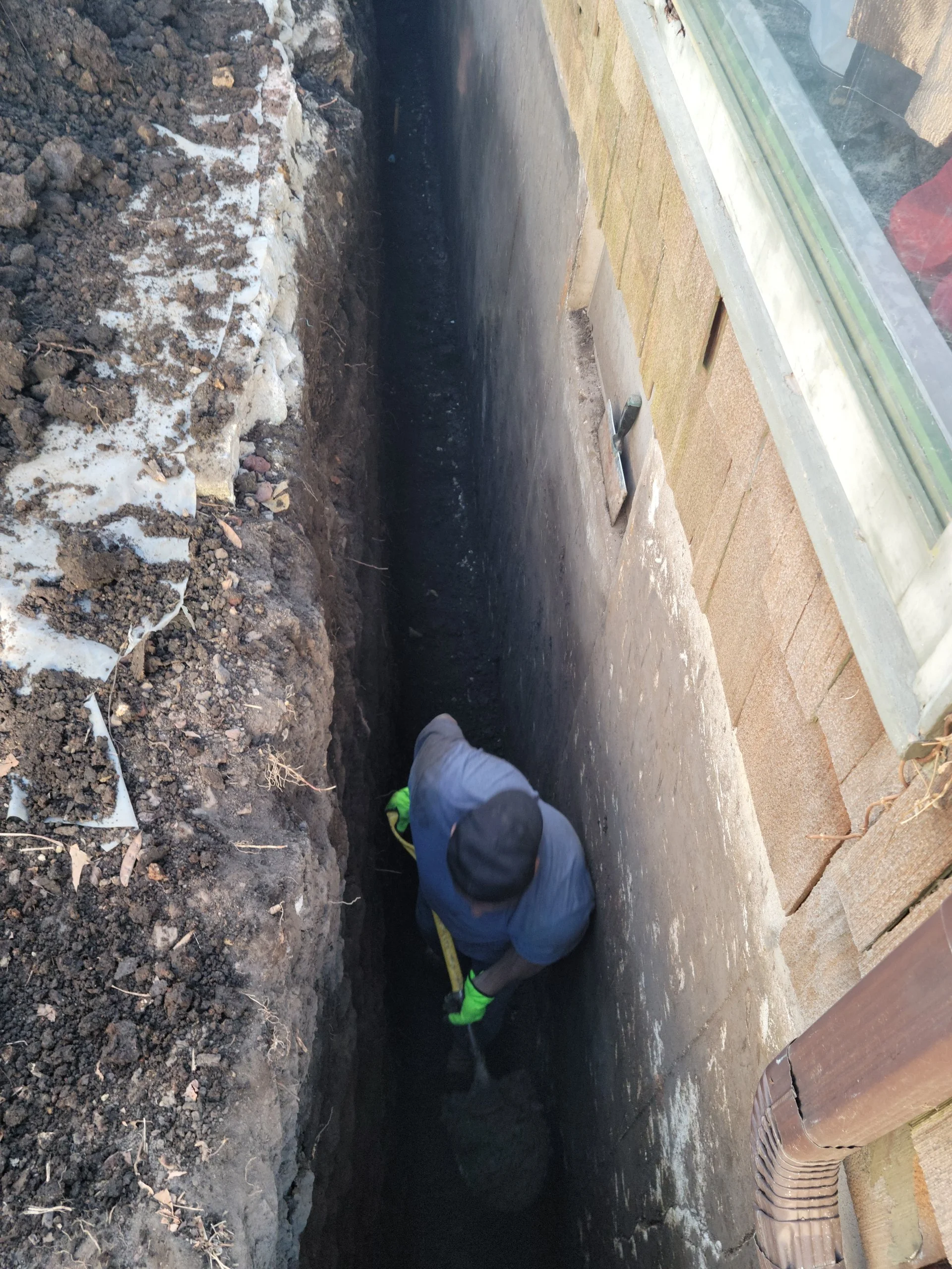 A person wearing a hooded jacket and green gloves working in a deep trench next to a house foundation.