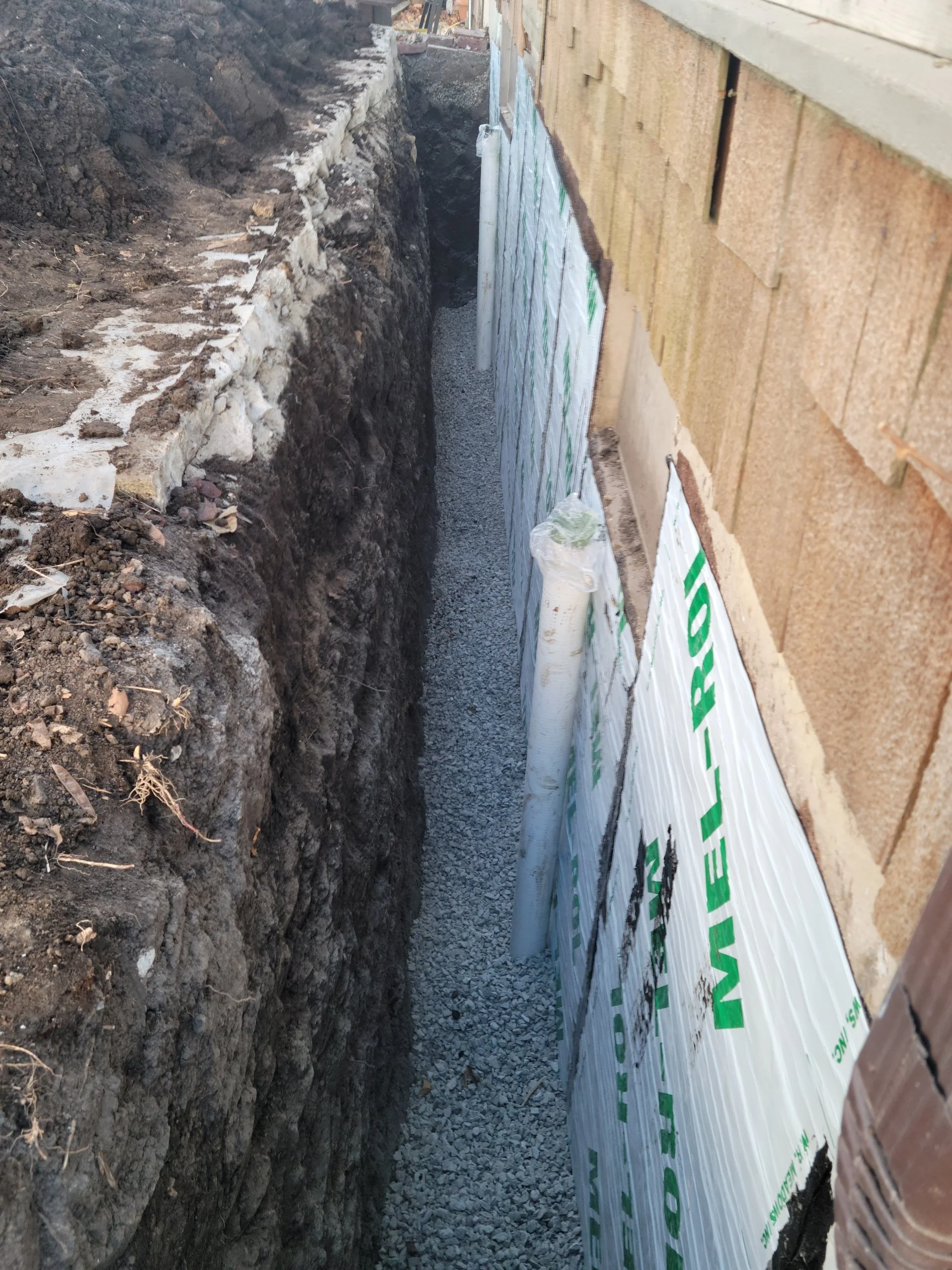 Construction site trench with gravel and pipes along the side of a building wall, using insulation materials.