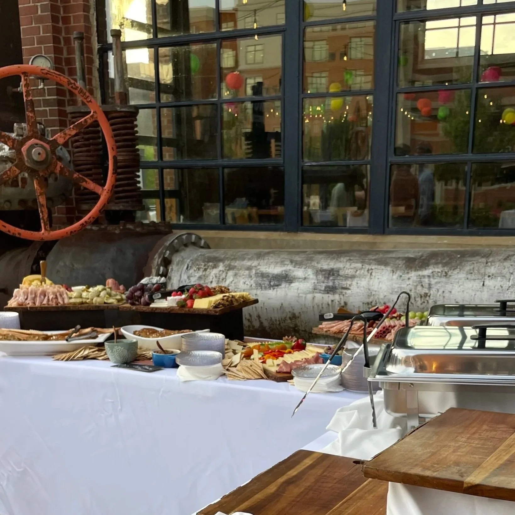 Buffet table with various dishes and snacks, set up outdoors in front of a large window reflecting the street outside, with a rustic pipe and wheel decoration on the brick wall behind. The setup suggests a casual or semi-formal event.