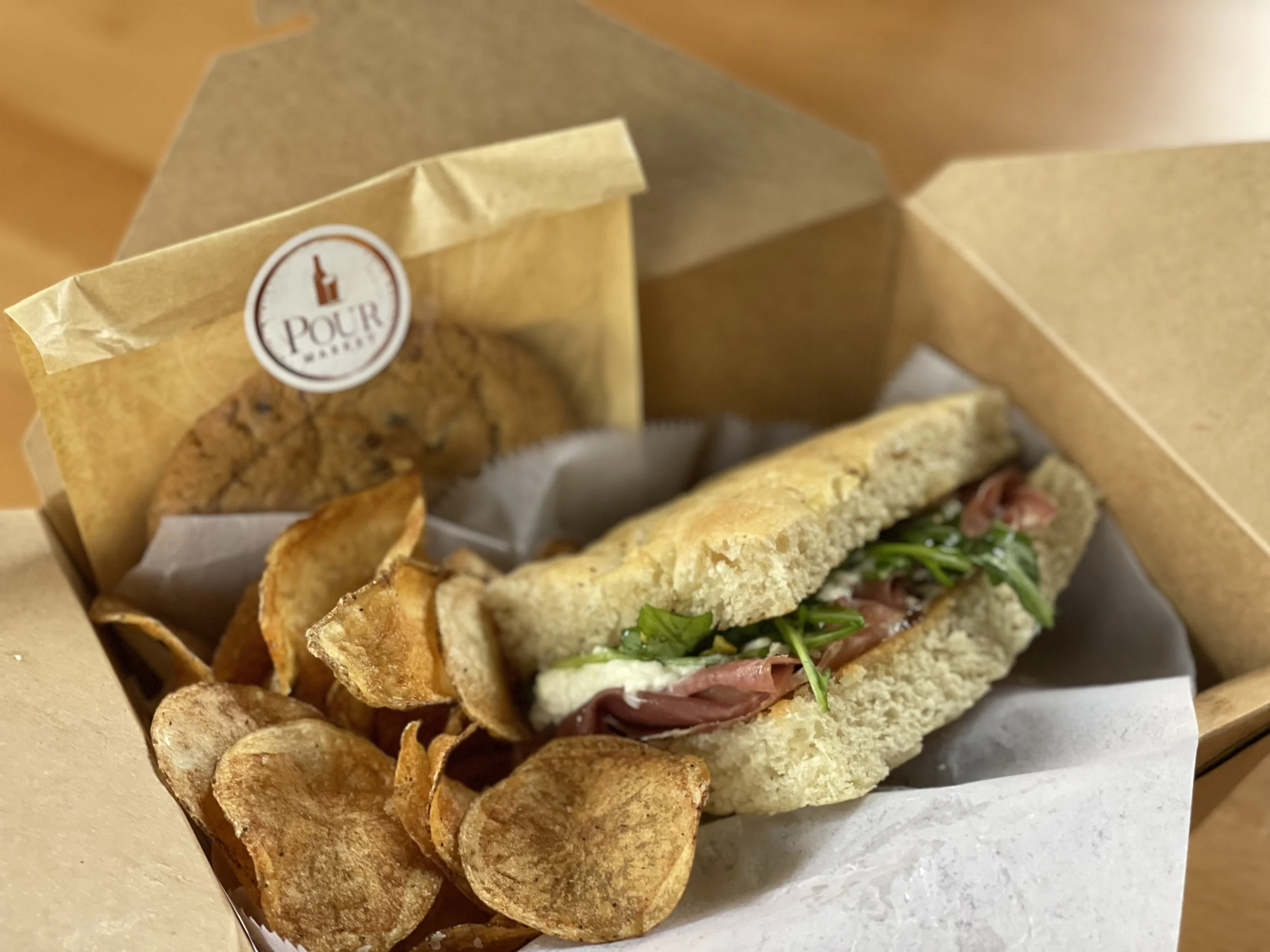 Sandwich with greens and meat, potato chips, and a cookie in a brown cardboard box.