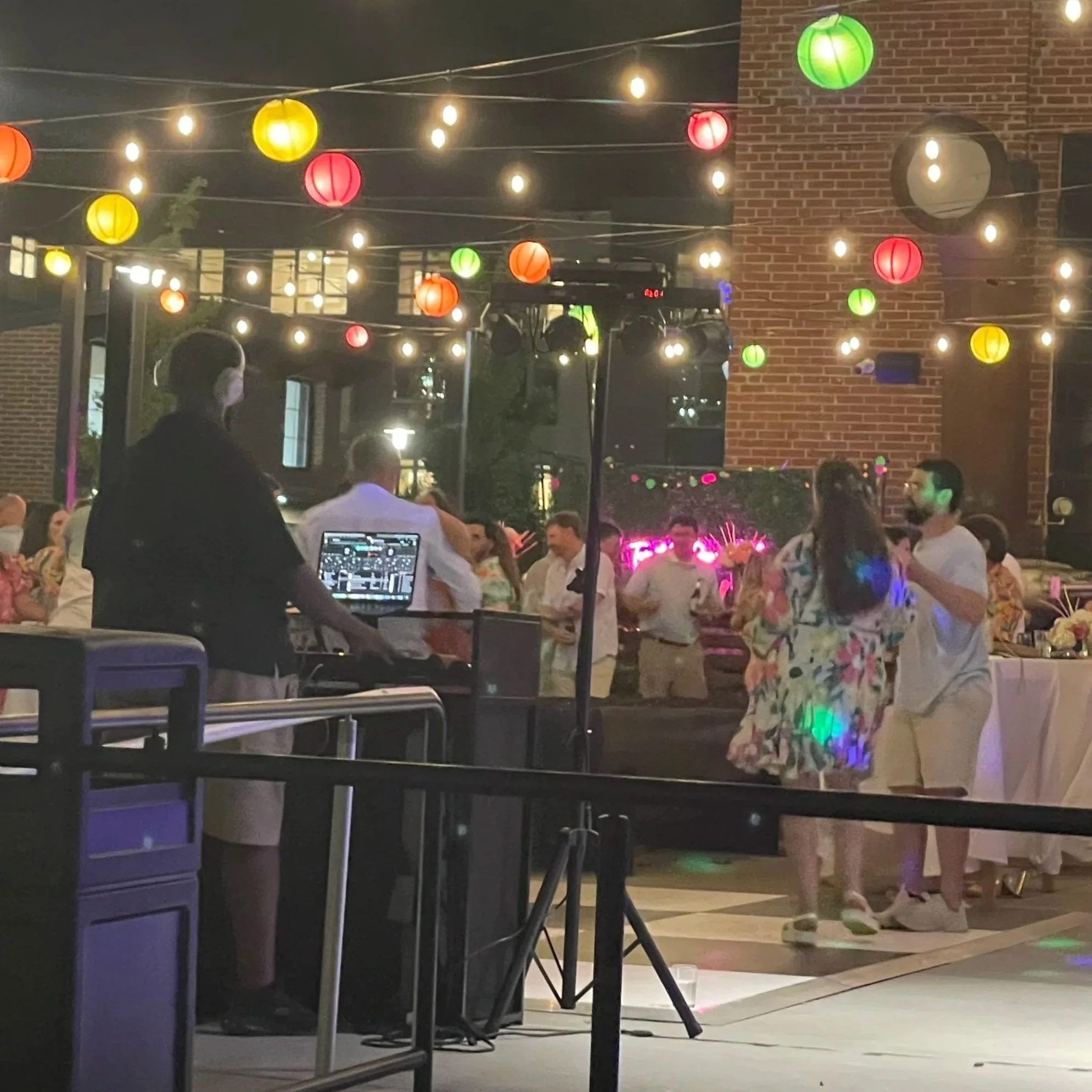Night scene of an outdoor party with colorful hanging paper lanterns and string lights, a brick building wall, and people enjoying the event.