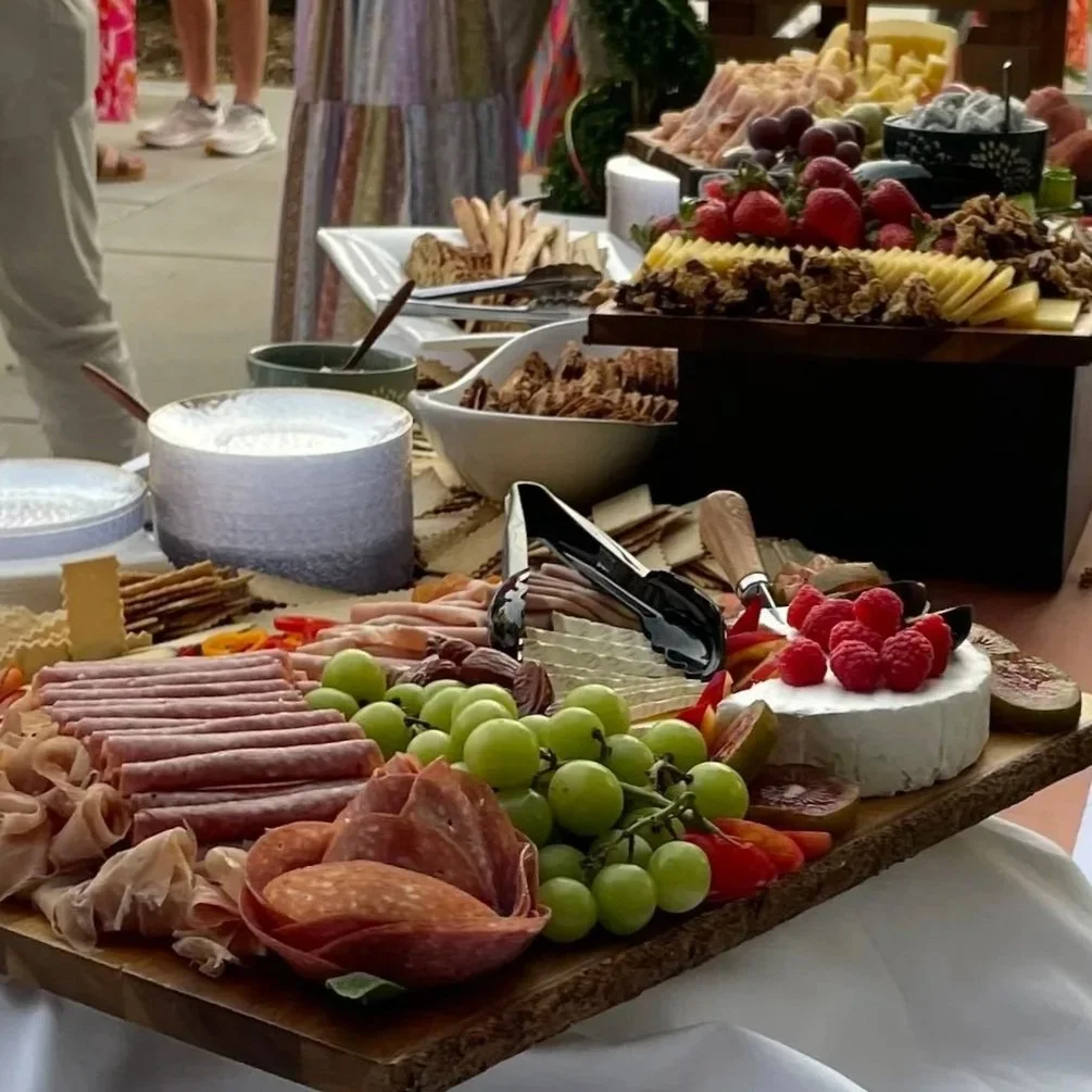 A charcuterie and cheese platter with grapes, raspberries, meats, cheeses, and crackers, alongside bowls of dips and other food items on a table.