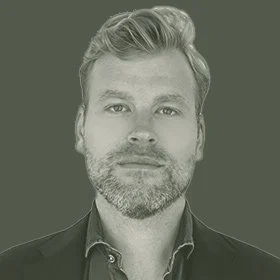 Black and white portrait of a man with short, styled hair and a beard, wearing a dark jacket and shirt, looking directly at the camera.
