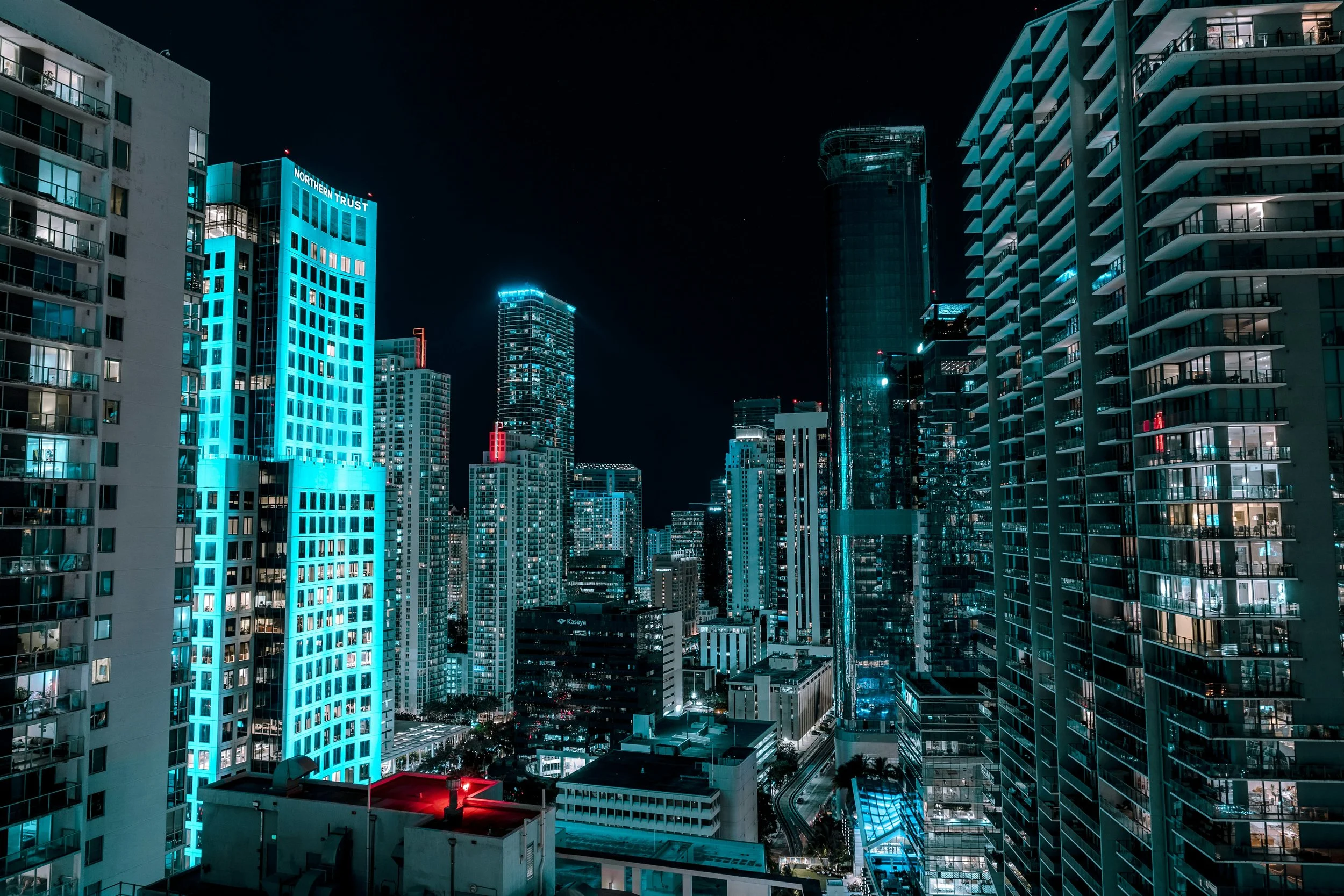 Night view of a city skyline with illuminated high-rise buildings and skyscrapers, some with blue and red lighting, and streets with light trails.
