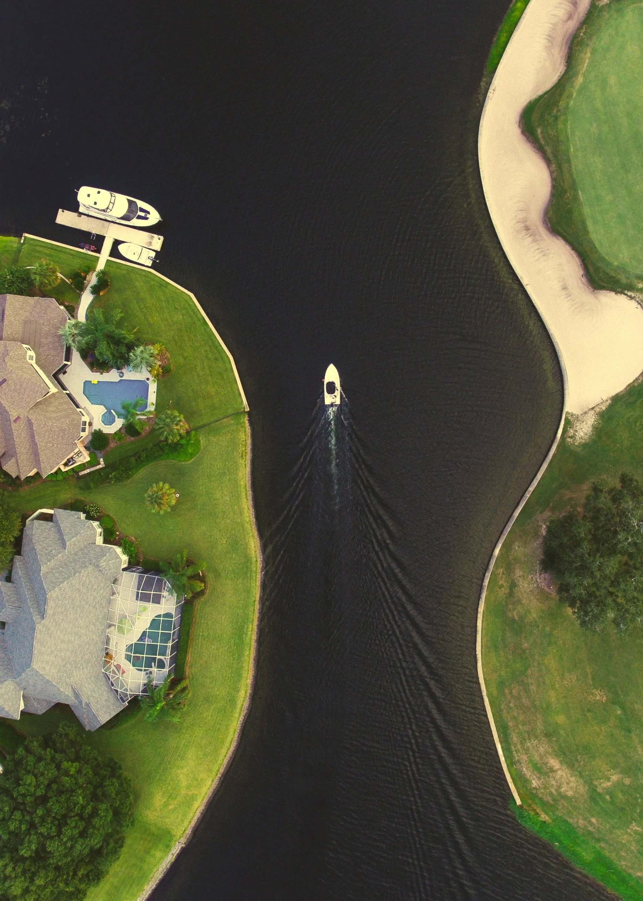 An aerial view of a boat moving through a canal between two residential neighborhoods with green lawns, trees, and houses, some with swimming pools, and a sandy beach area on the right side.