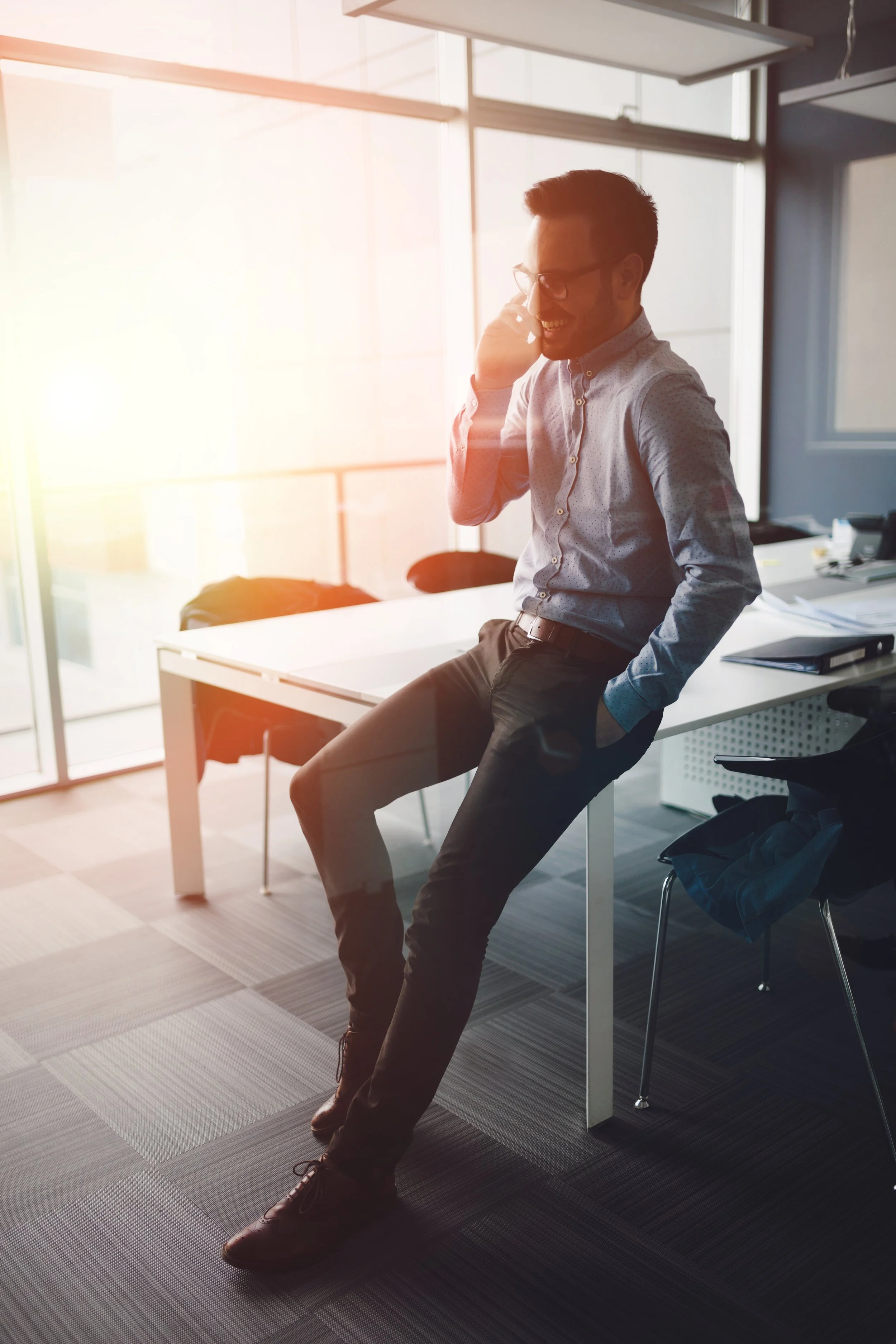 A man with glasses and a beard, dressed in a light patterned shirt and dark pants, is talking on the phone while leaning against a white desk in an office with large windows and bright sunlight.