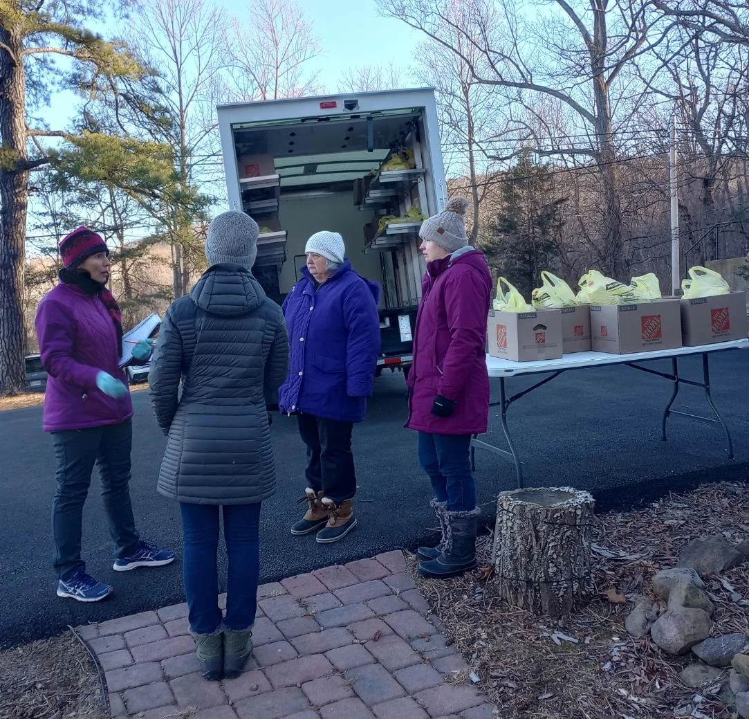 Four women wearing winter clothing and hats standing outdoors near a truck and a folding table with boxes of supplies, possibly distributing goods or food.