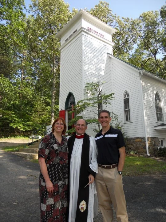 Three people stand outside a white church with tall windows, green trees in the background, and steps leading up to the entrance.