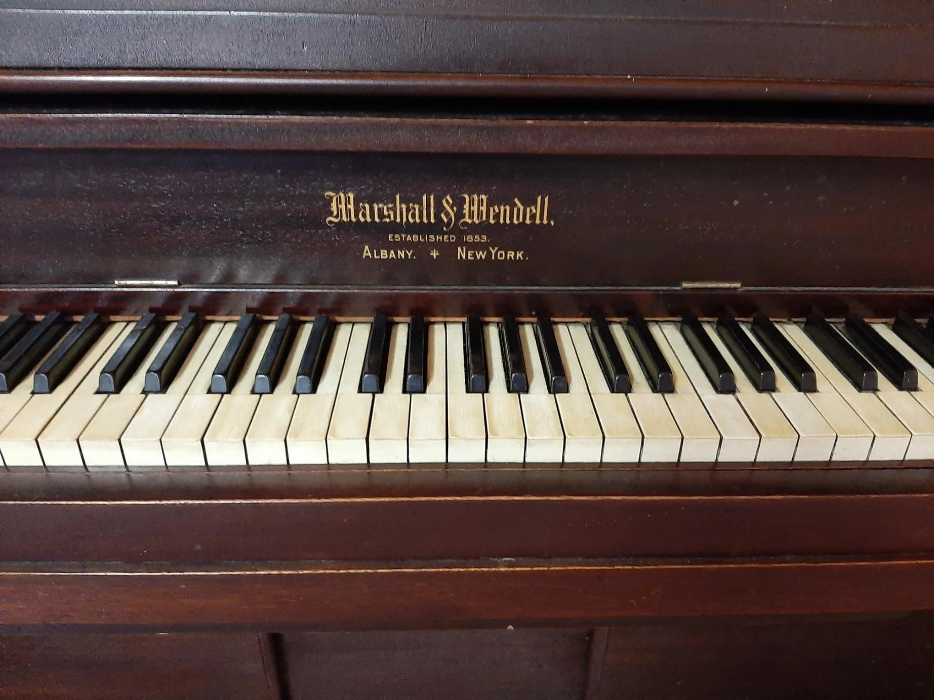 An antique upright piano with a wooden case, opened keyboard showing black and white keys, and labeled 'Marshall S. Wendell, Established 1853, Albany, New York'.