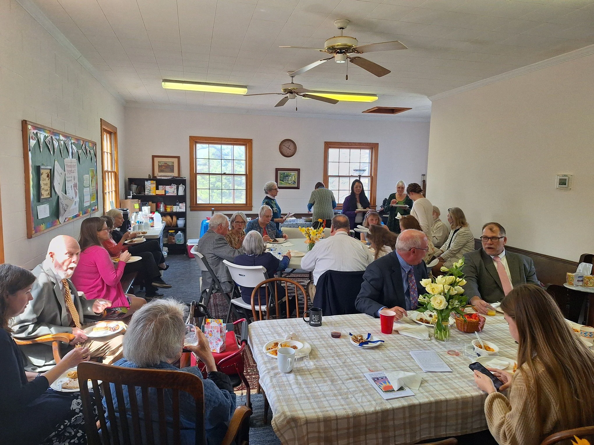A large gathering of mostly elderly people seated at tables in a community room, sharing a meal, with some standing and serving food near the back. The room has two large windows, ceiling fans, and a bulletin board on the wall.