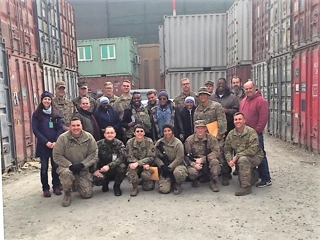 Group of soldiers and civilians standing and kneeling among shipping containers outdoors.