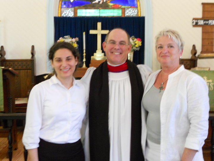 A pastor standing with two women inside a church. The pastor is wearing a white robe with a red collar and a black stole. The woman on the left is wearing a white blouse and black skirt, and the woman on the right is wearing a white blazer. They are 