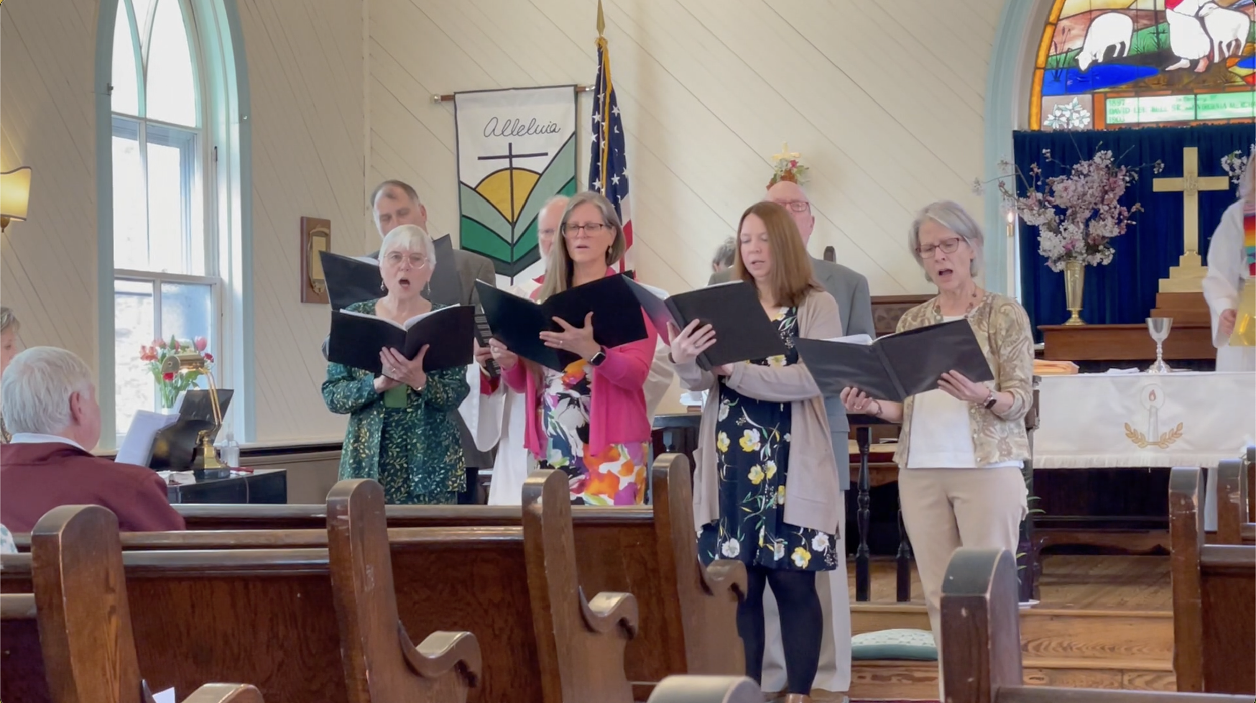 Church choir performing during service inside a church with stained glass windows, wooden pews, and religious symbols.