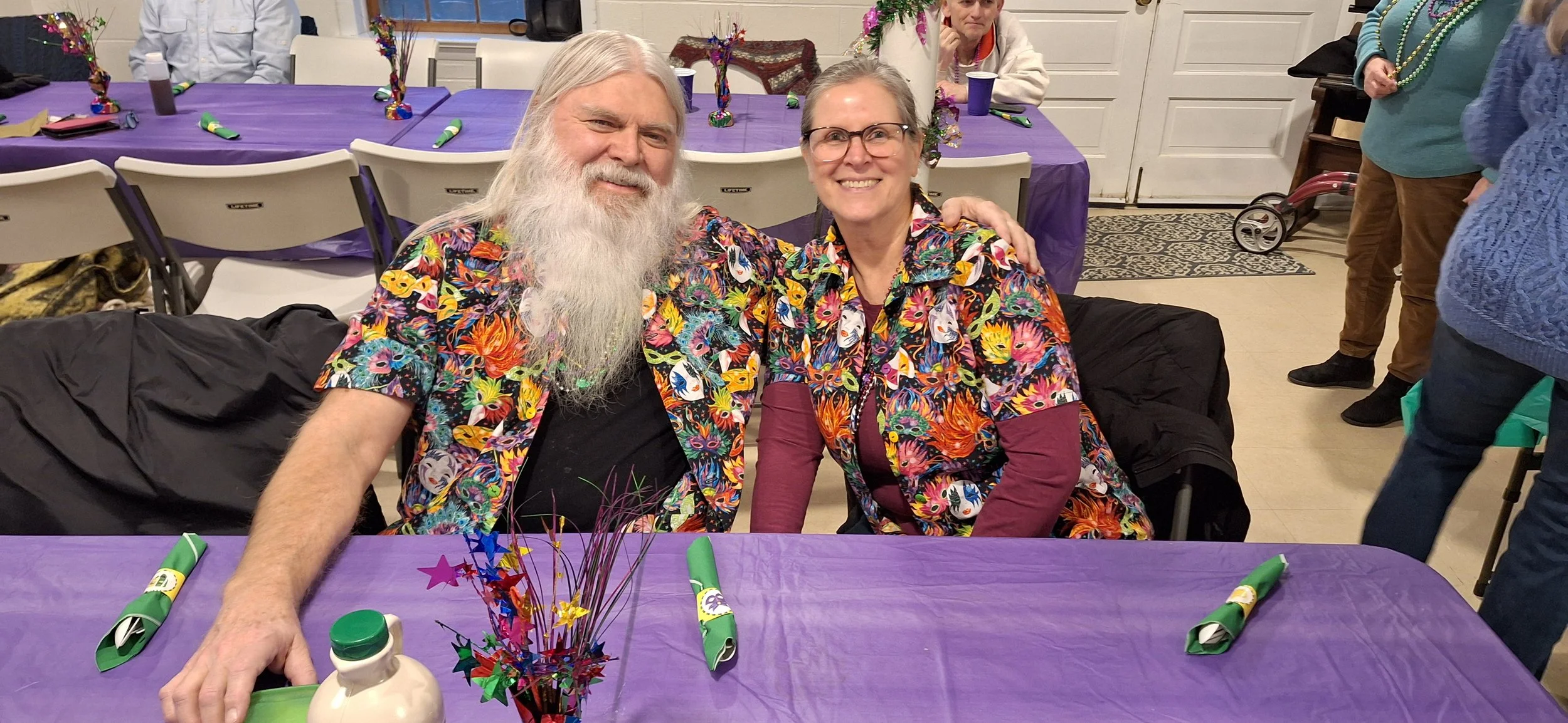 A man and woman sitting at a table, both wearing matching colorful, patterned shirts with skulls, flowers, and faces, smiling for the camera in a party setting with purple tablecloths and party decorations around.