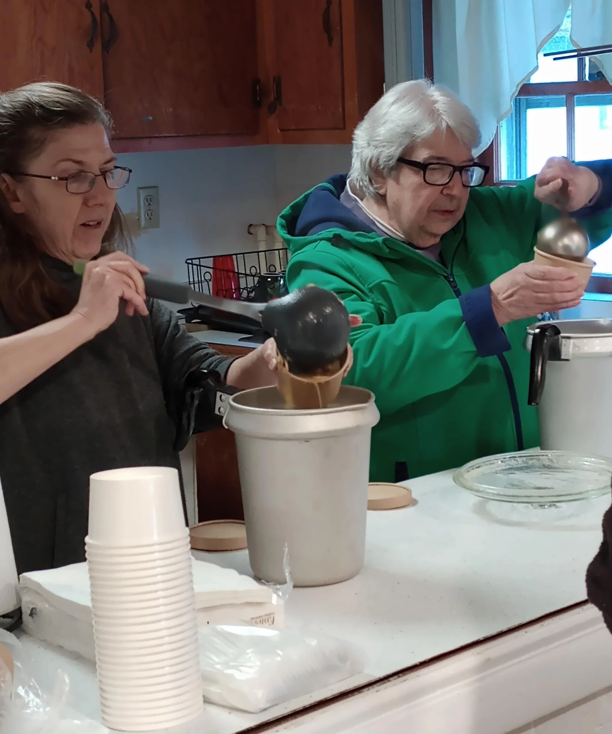 Two women pouring ice cream from scoops into cups in a kitchen