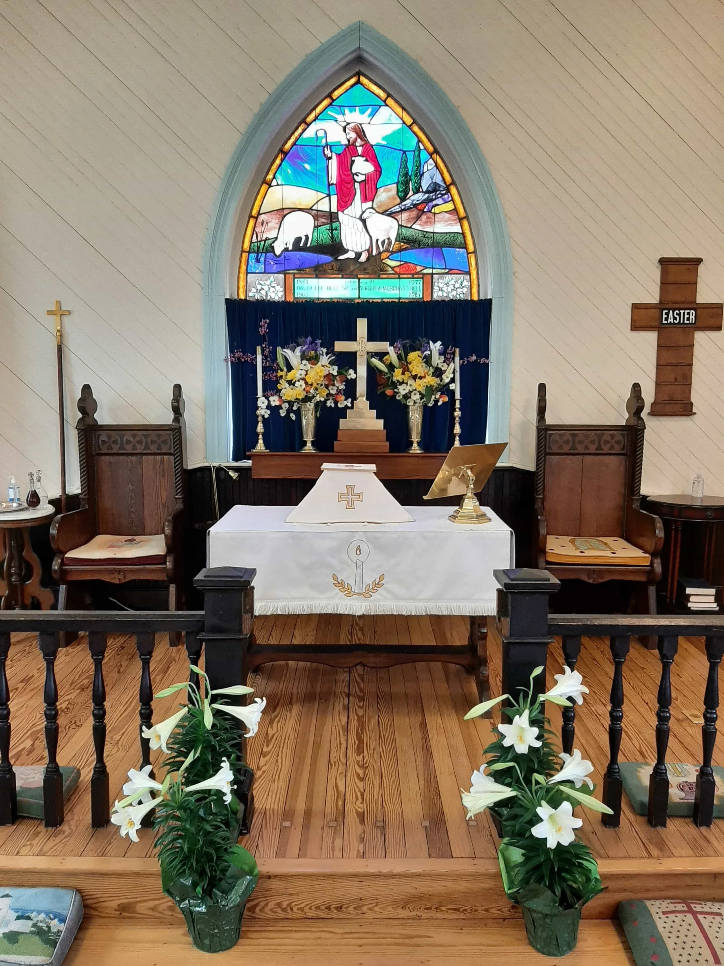 Inside a church, an altar decorated with flowers, a cross, and a stained glass window depicting Jesus with sheep, with lilies in front and wooden chairs surrounding it.