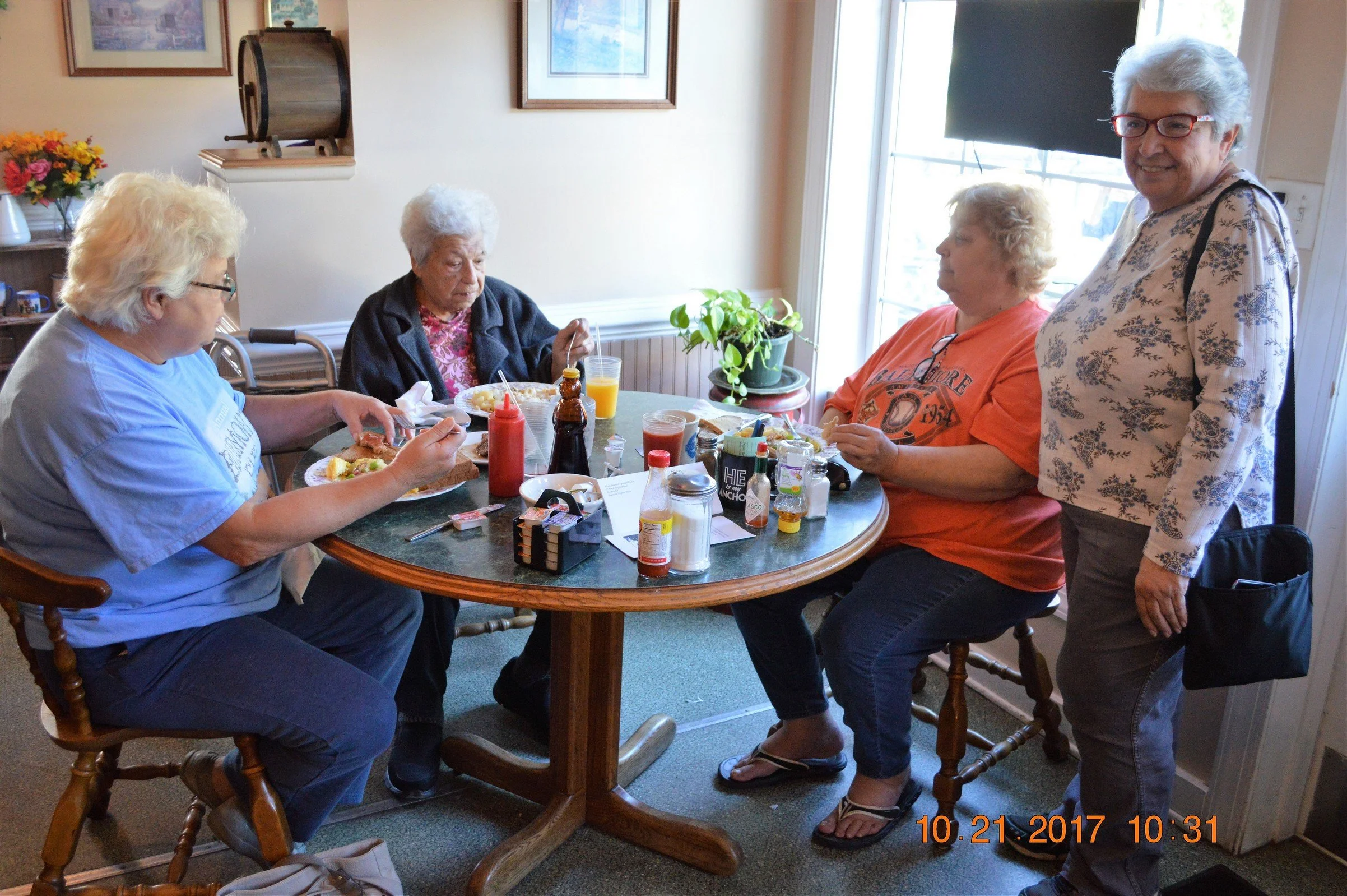 Five women gathered around a dining table in a kitchen, some eating and some talking, with condiments and drinks on the table, and a window letting in natural light.