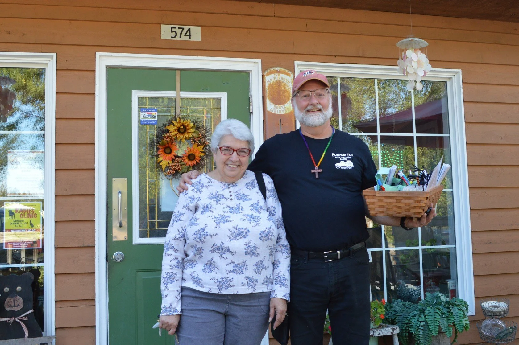A smiling elderly woman with white hair and glasses standing next to a smiling man with a beard, glasses, and a pink cap, outside a building with a green door and window. The man holds a basket filled with various items. The background includes a dec