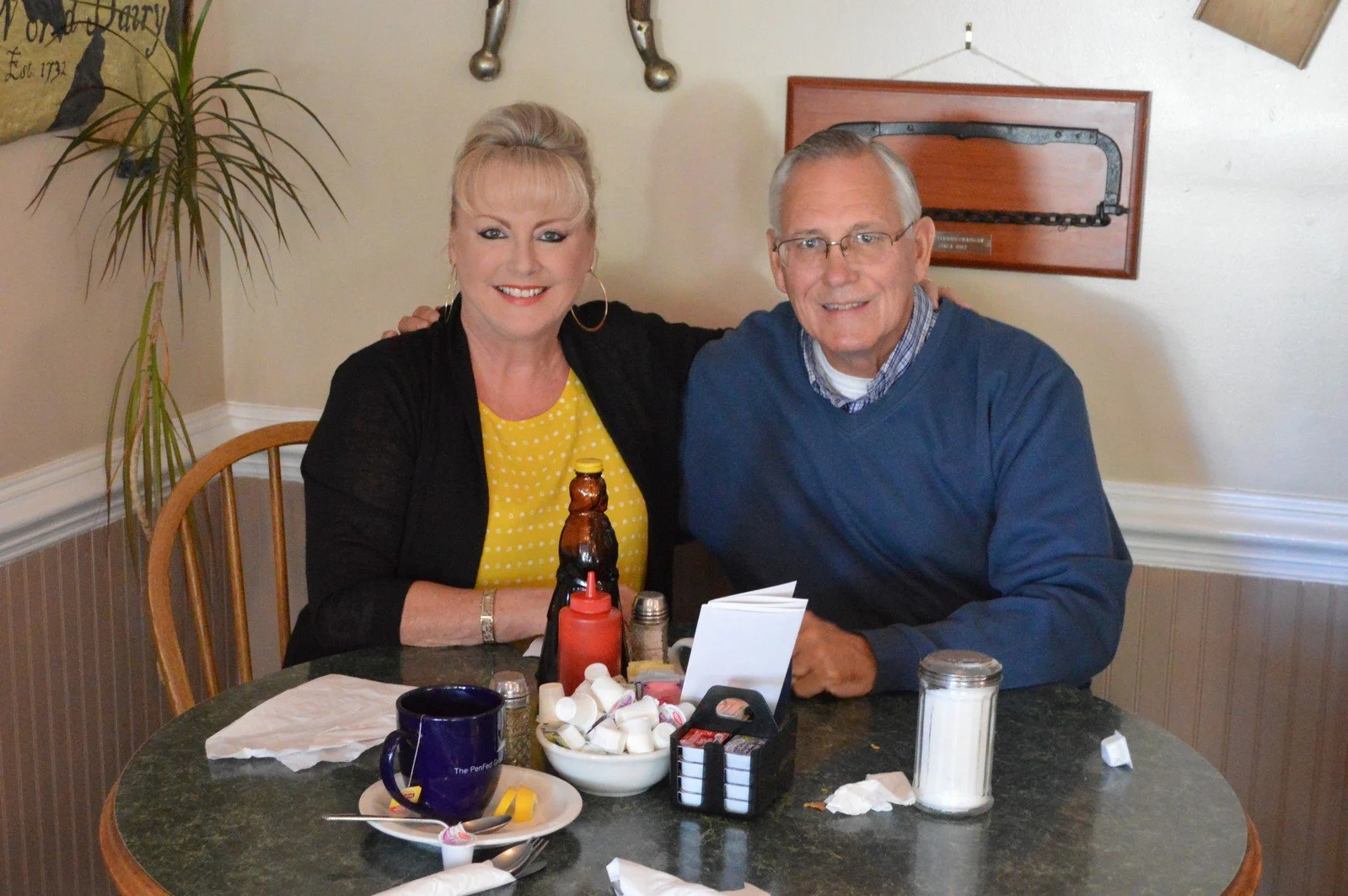A woman and a man sitting at a restaurant table, smiling. The woman wears a yellow polka dot shirt and black cardigan, and the man wears glasses and a blue sweater. The table has a bowl of sugar packets, condiments, coffee mug, and sugar dispenser.