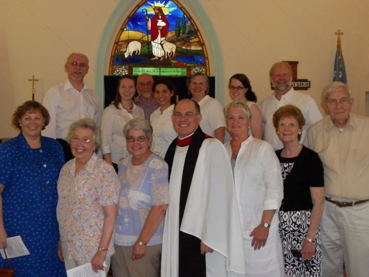 Group of people posing inside a church near a stained glass window depicting Jesus with sheep and the Good News Bible verse.