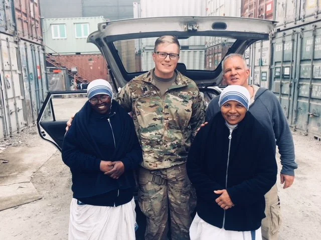 Four people standing in front of an open car trunk in a shipping yard, including two women in black jackets and headscarves, a man in camouflage military uniform, and another man in a gray hoodie.