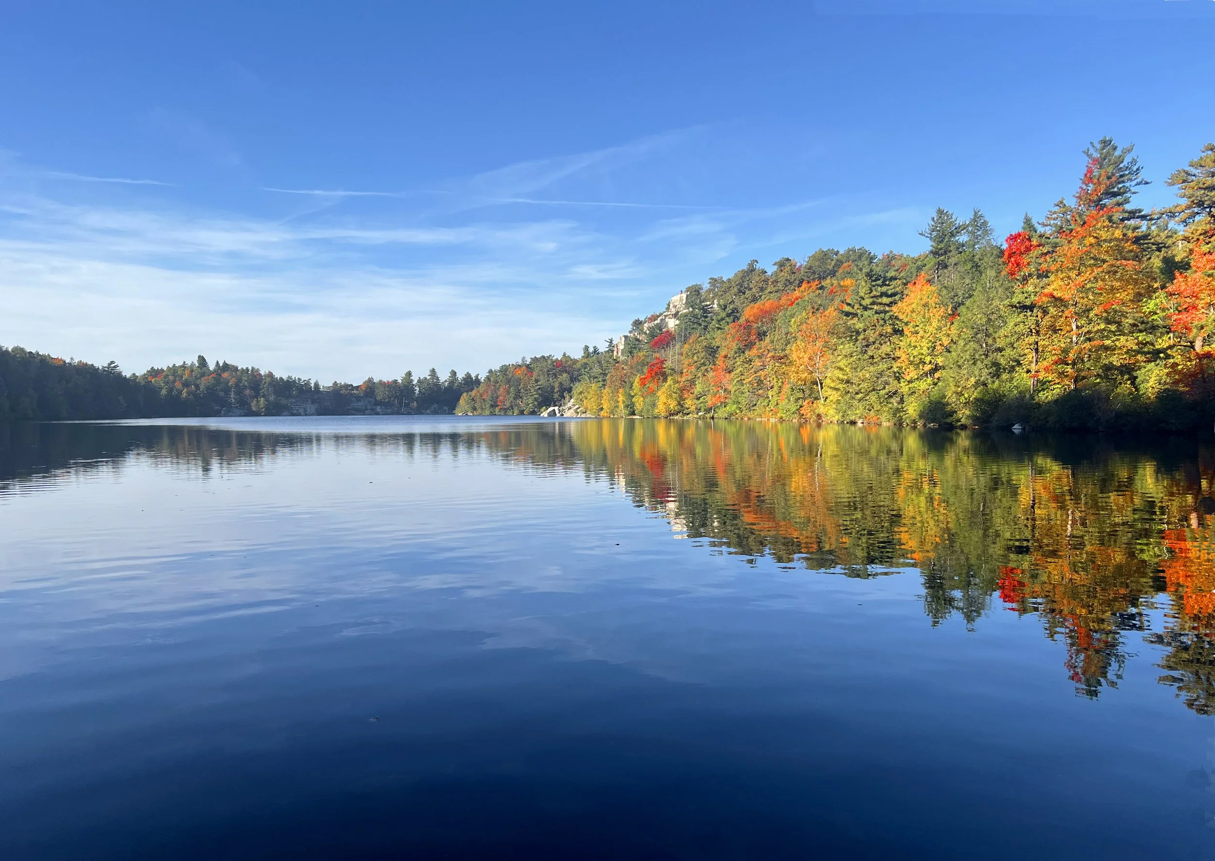 Calm lake with reflections of autumn-colored trees along the shoreline under a clear blue sky.