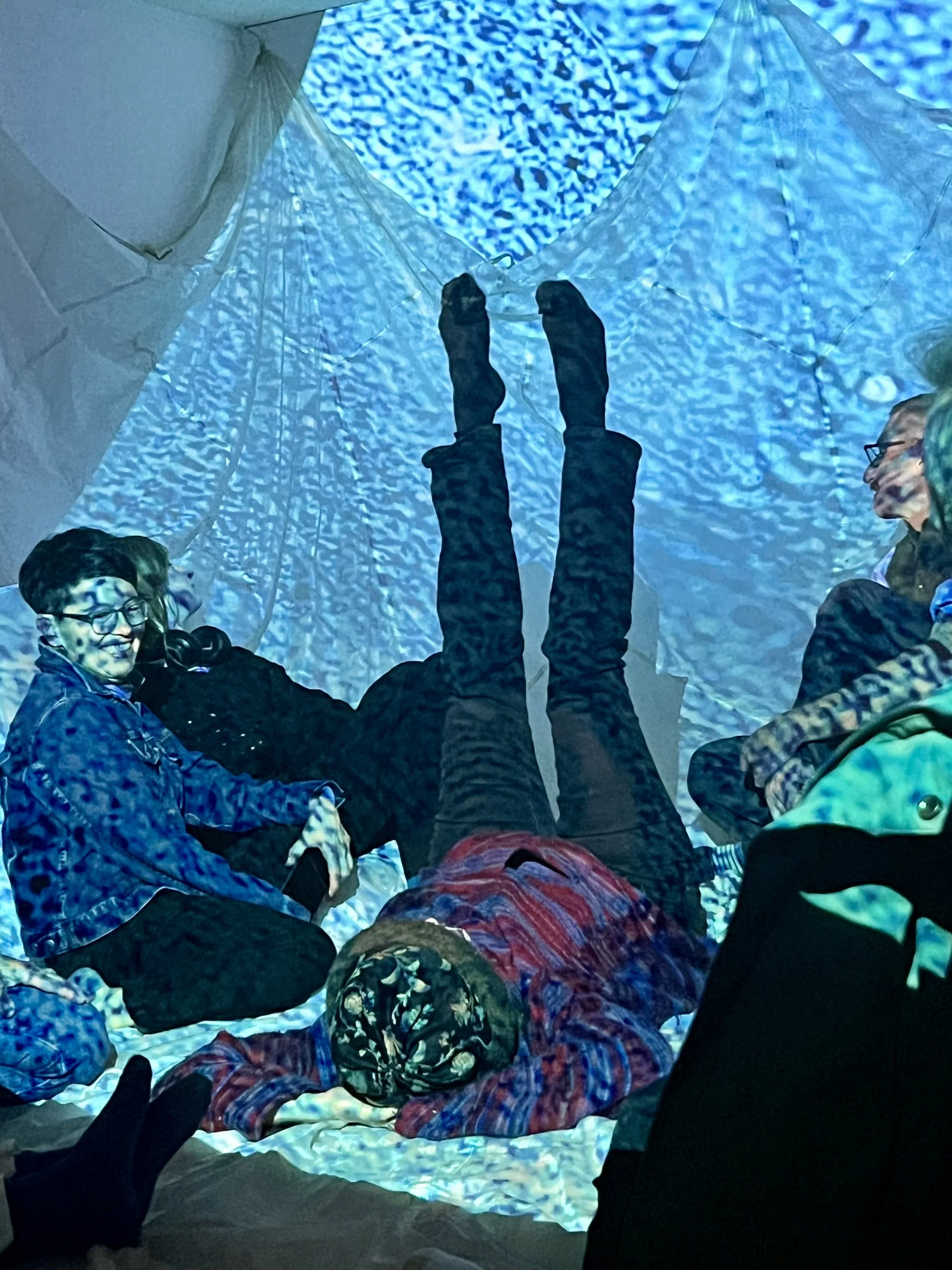 People sitting and lying inside an art installation including a white tent with textured blue fabric, some smiling and others relaxing, with a person lying on the ground wearing a patterned headwrap and glasses.