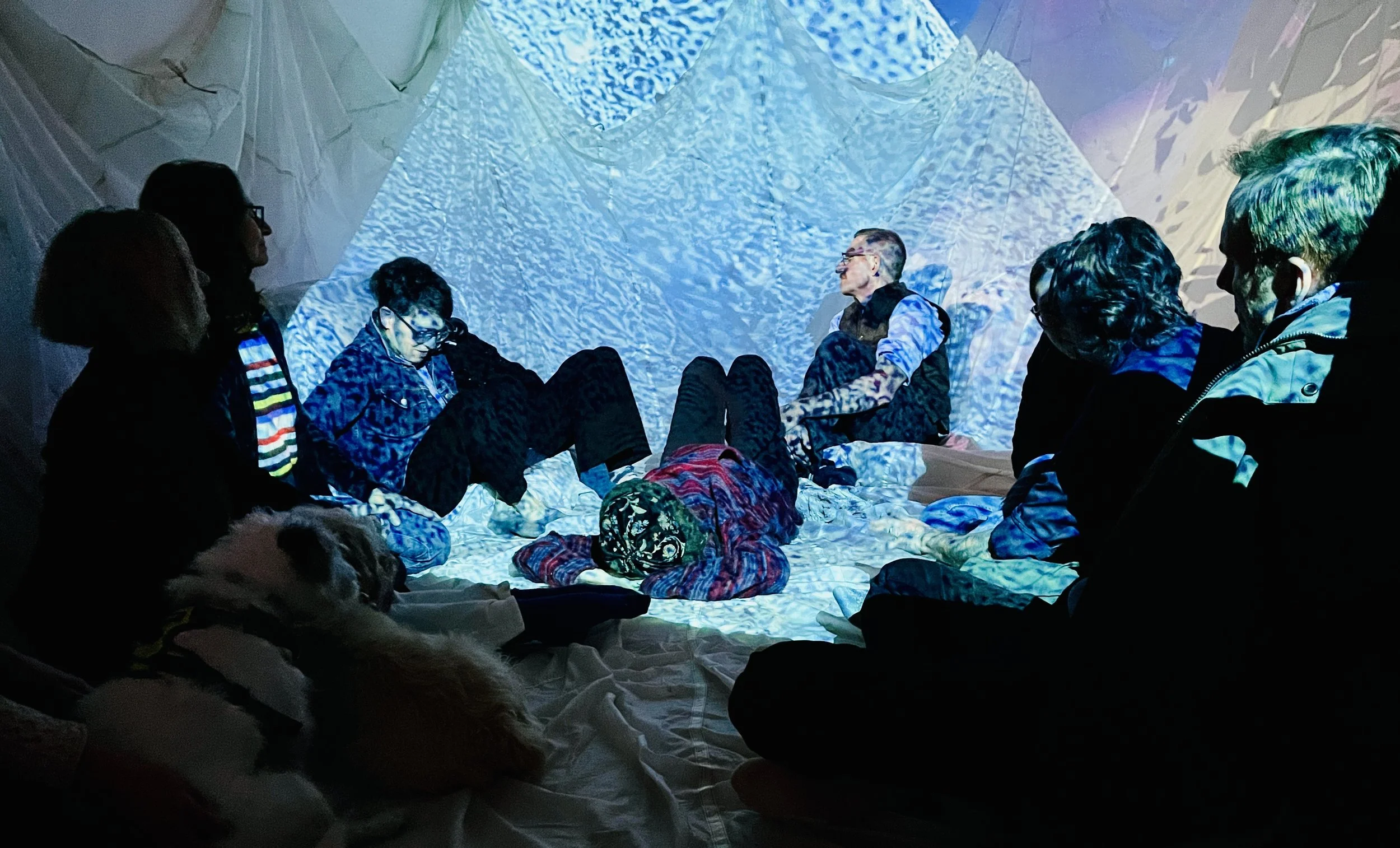 People sitting inside a tent with projected blue and white abstract patterns on the walls and ceiling, some with glasses and one with a dog, engaged in conversation or listening.
