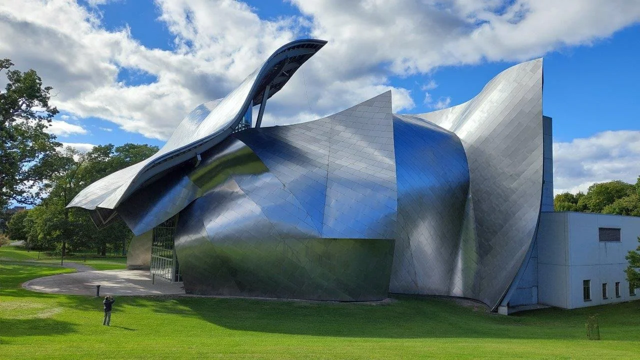 The Guggenheim Museum in New York City, featuring an avant-garde, metallic, curvy architectural design with reflective surfaces, surrounded by green grass and trees under a partly cloudy sky.
