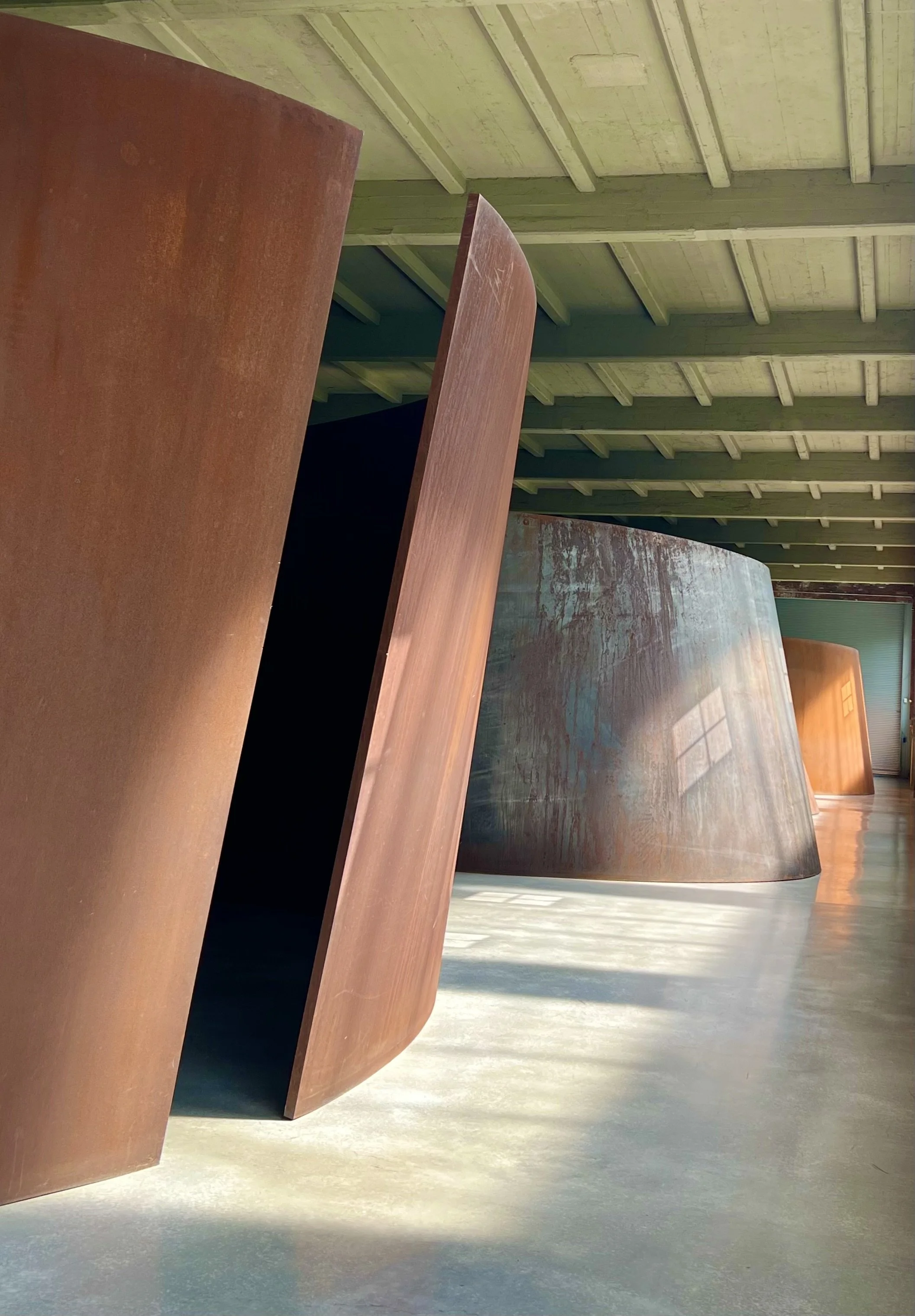 Large rusted metal sculptures with curved shapes indoors, sunlight casting shadows on the smooth concrete floor at Dia Beacon.