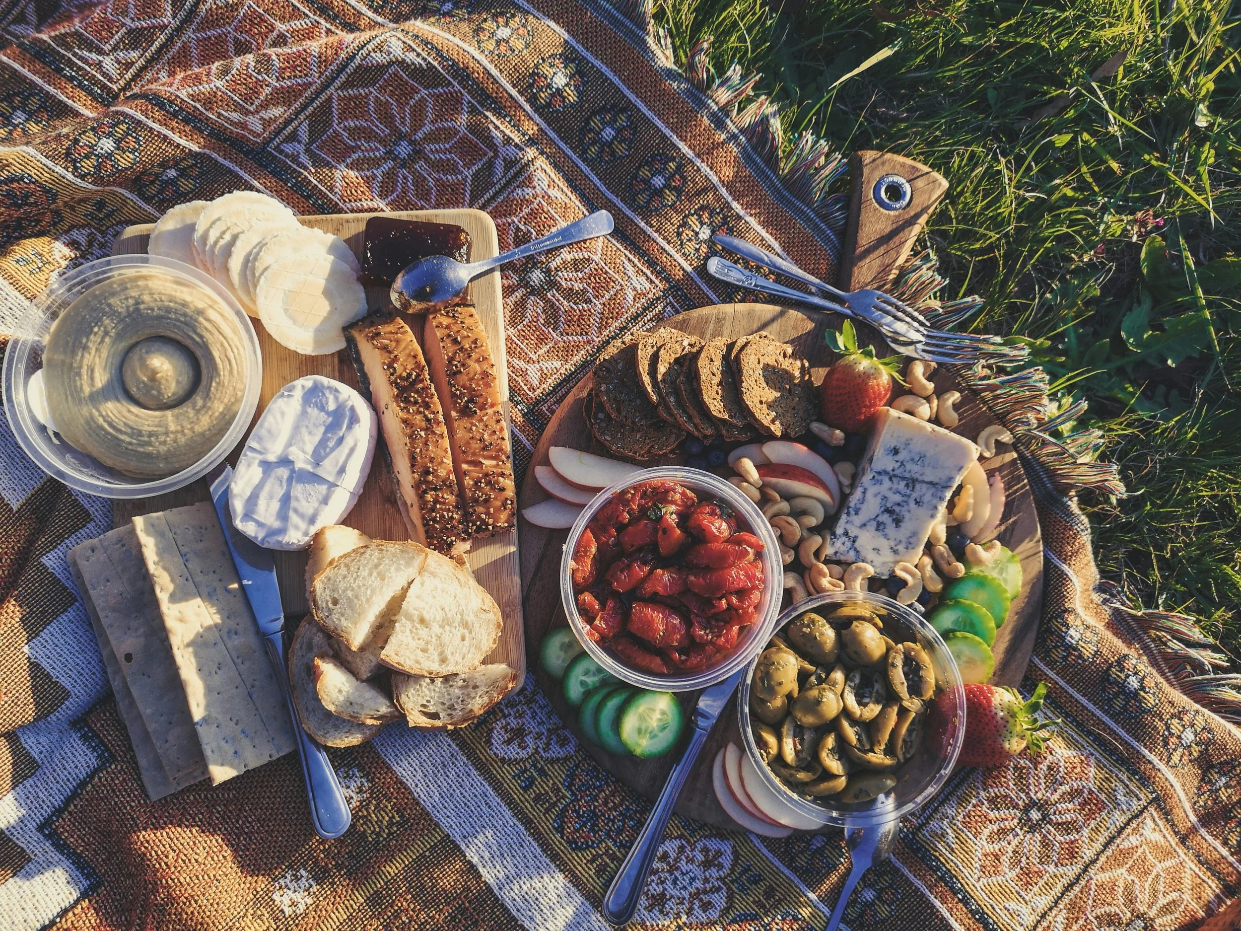 A picnic spread on a patterned blanket in a grassy outdoor setting, featuring cheese, bread, sliced vegetables, strawberries, olives, nuts, and small plates of spread and jam.