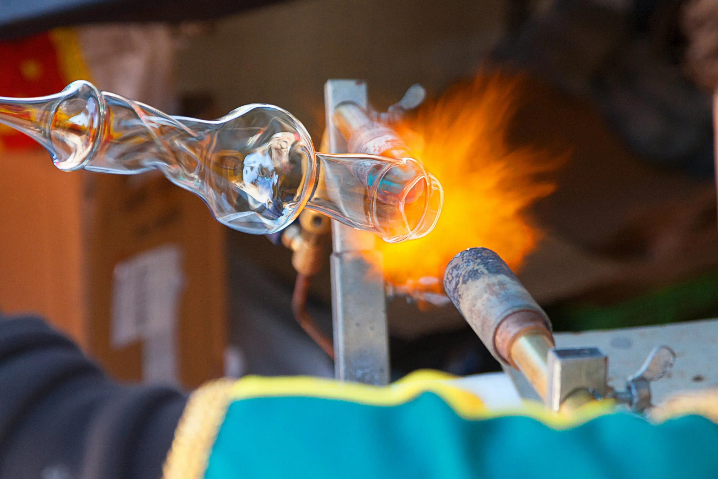 Glassblower shaping molten glass in a furnace, with bright orange flames, in a glassmaking studio.