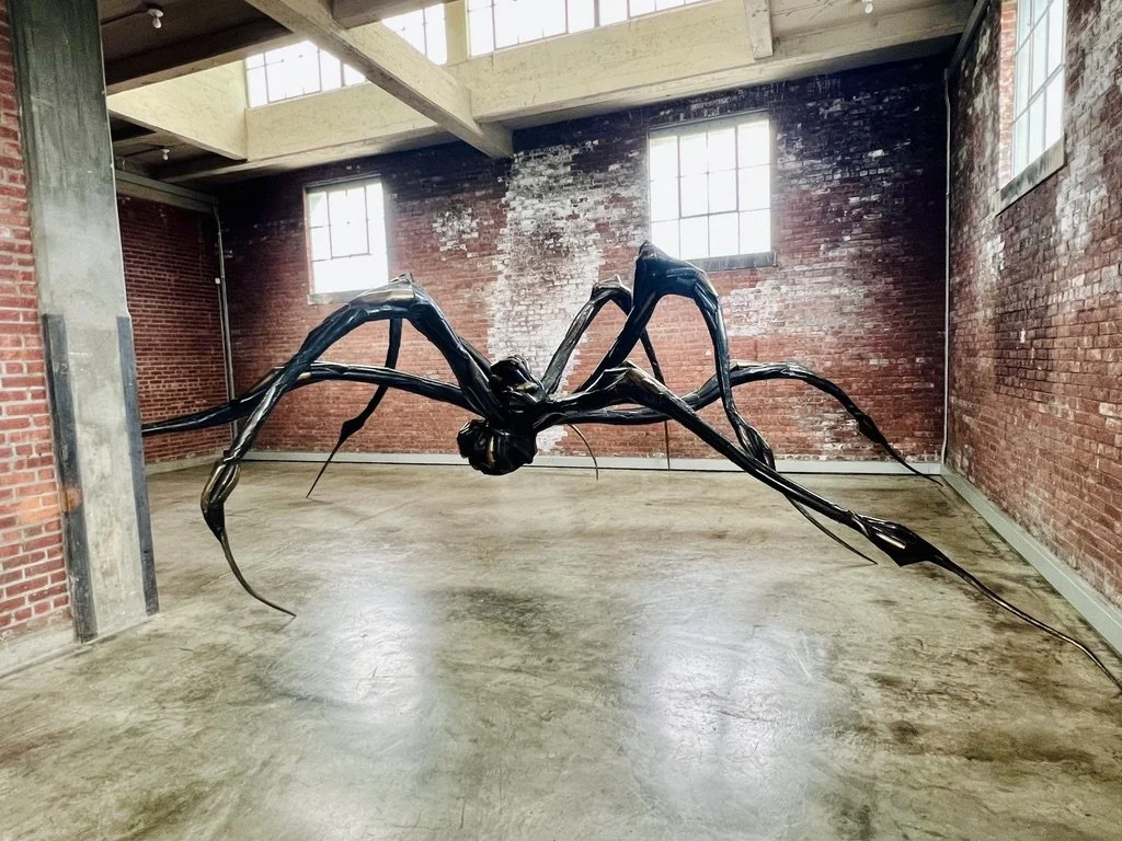 A large black abstract sculpture resembling a spider with elongated legs, situated in an industrial-style room with exposed brick walls and large windows at Dia Beacon.