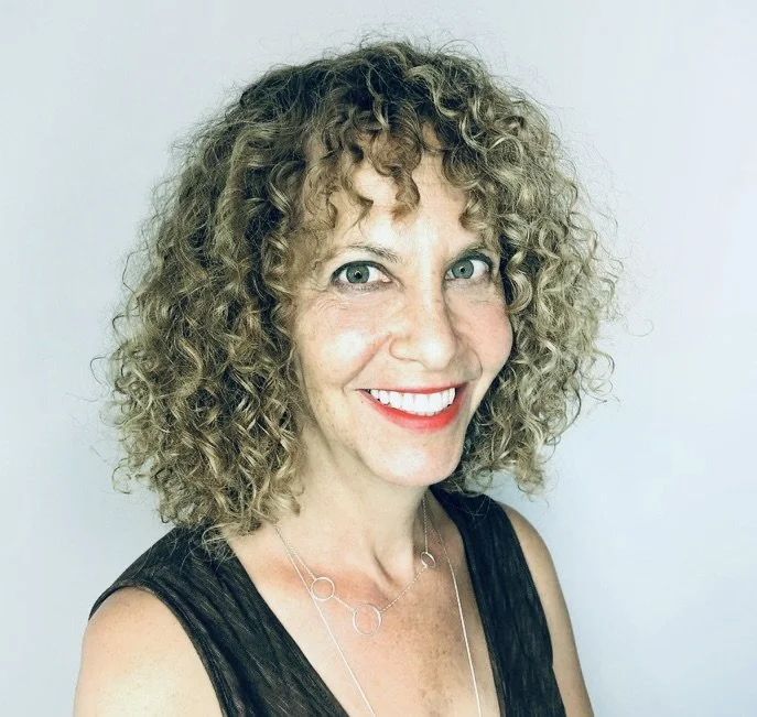 Artist with curly blonde hair smiling at the camera, wearing a black sleeveless top and a silver necklace.