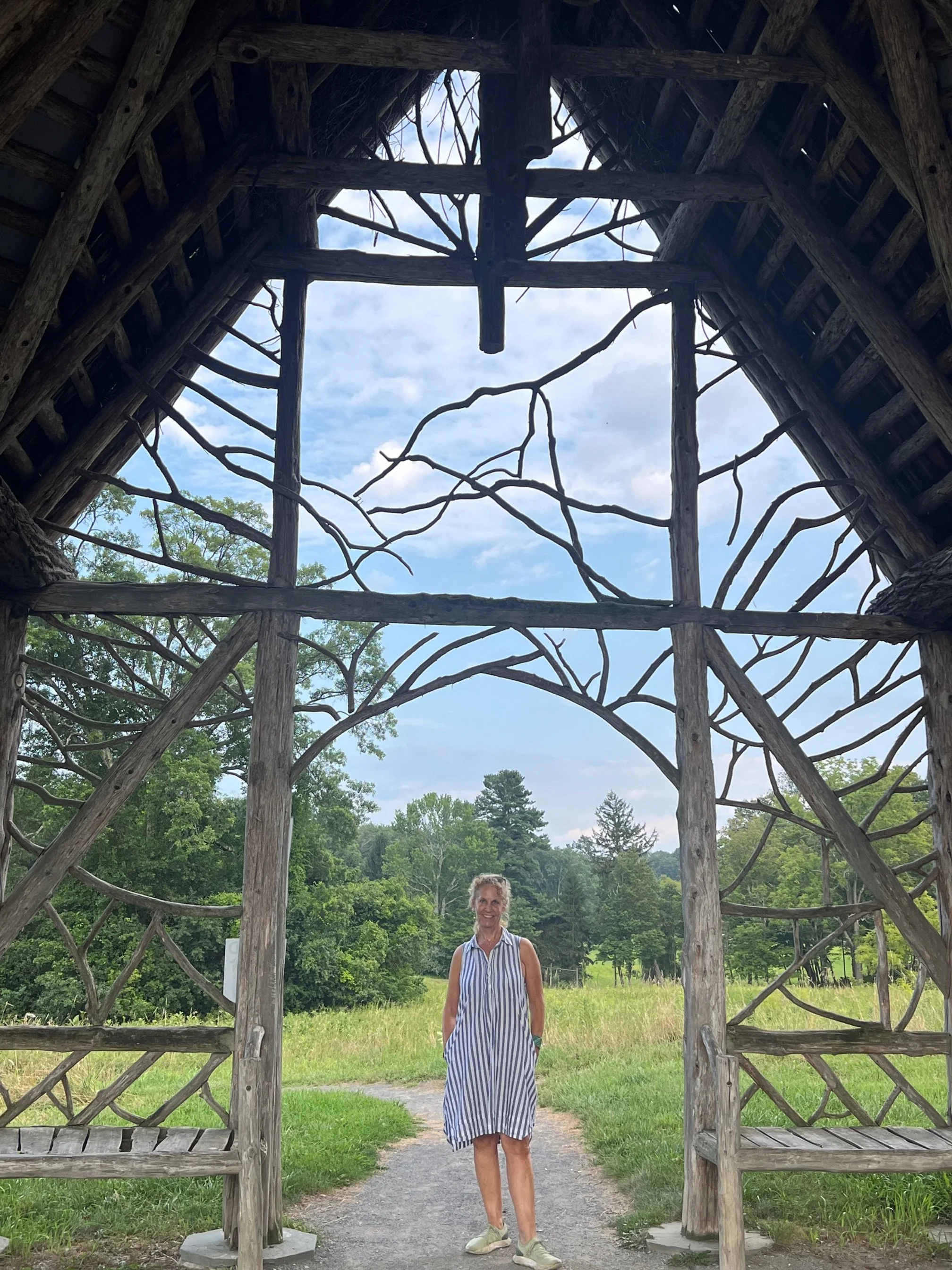 A woman in a striped dress standing on a gravel path beneath a wooden structure with intertwined branches and open sky at Poet's Walk Hudson Valley.
