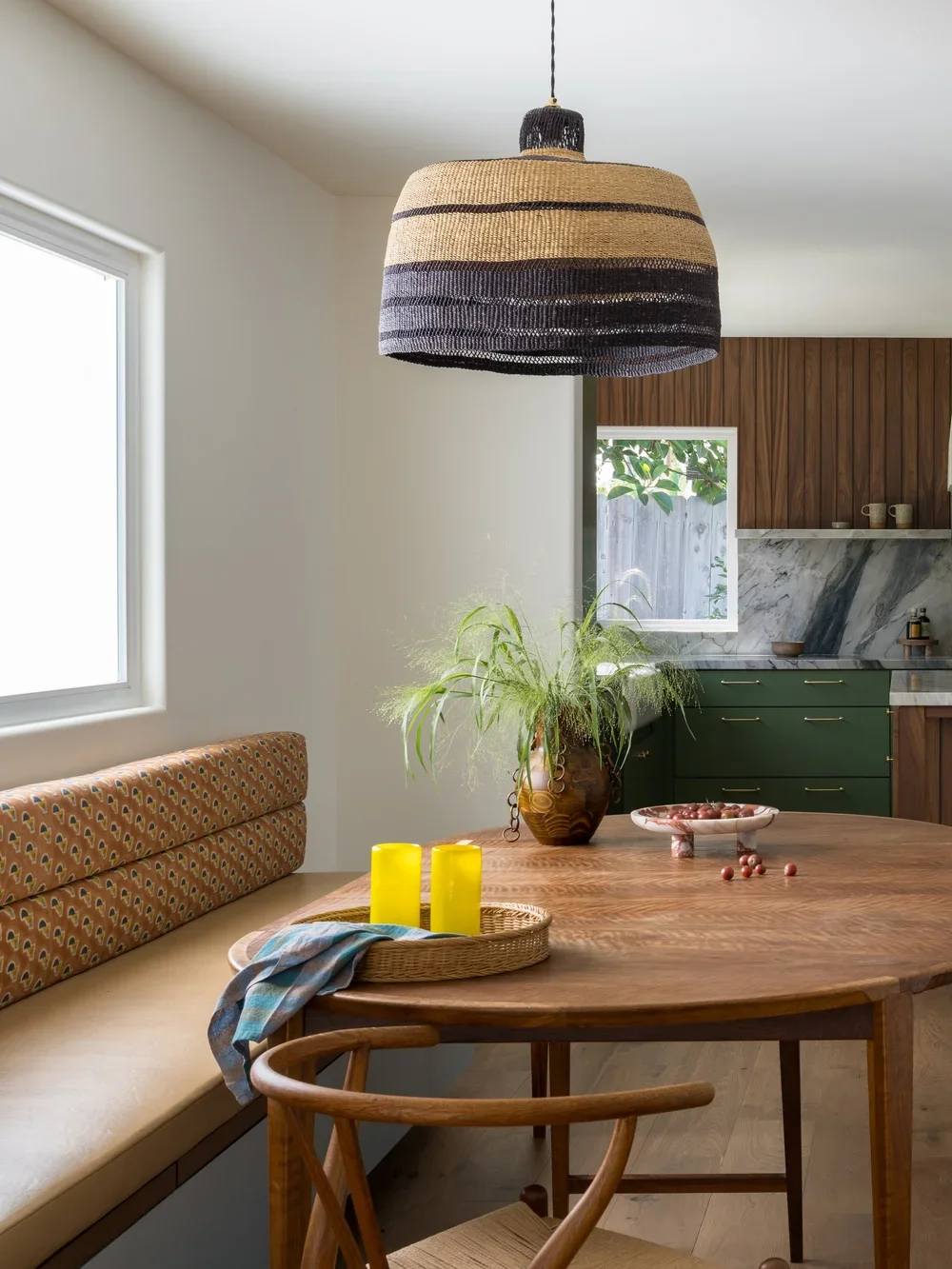 A dining room with a wooden table and a woven pendant light hanging above it. The table has yellow candles, a basket with a towel, and a pink dish with small round objects. There's a built-in bench with patterned fabric and a large vase with green plants. The background shows part of a kitchen with green cabinets, marble countertops, and wooden wall panels.
