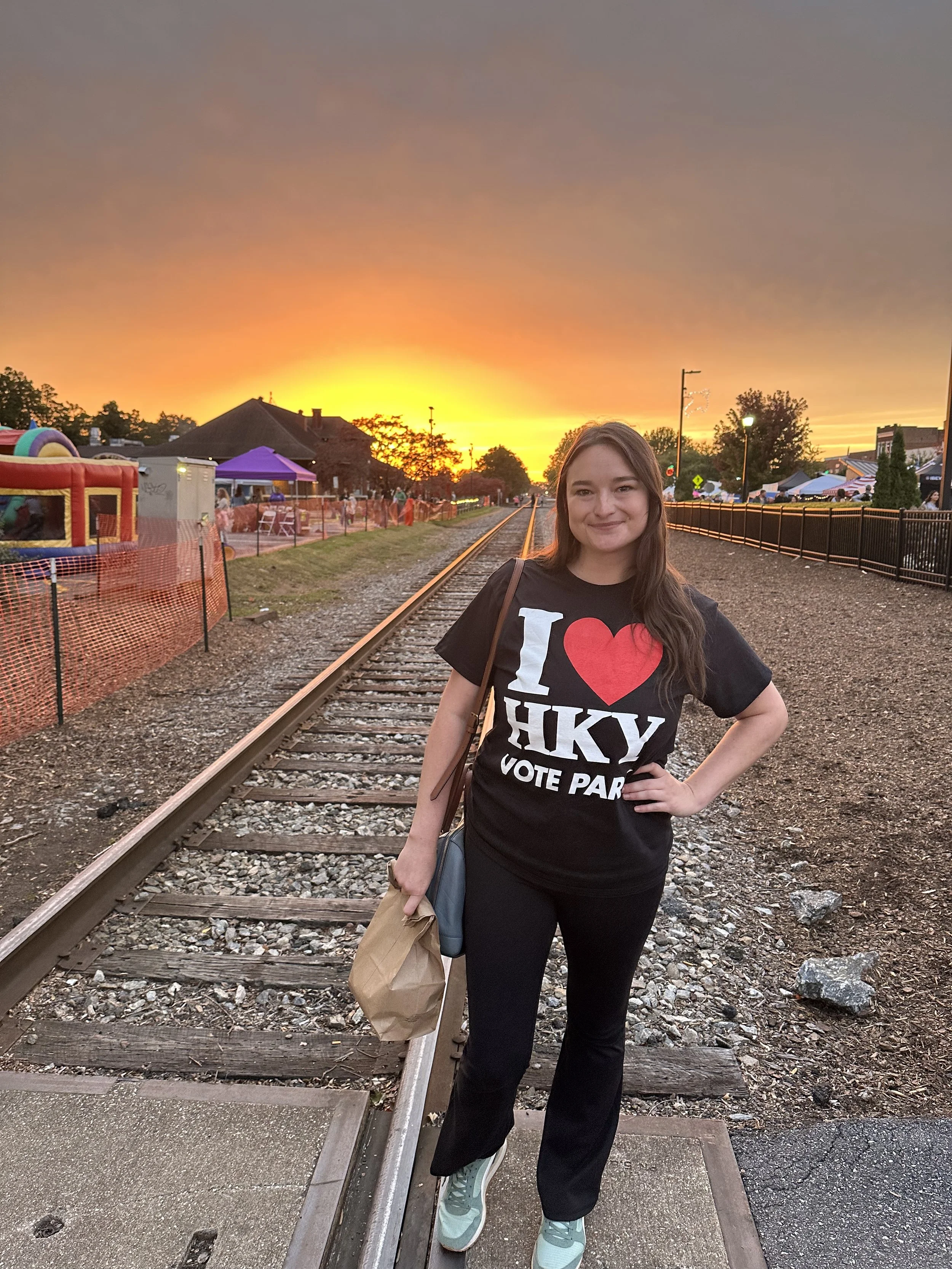 Park standing on railroad tracks at sunset, wearing a black t-shirt that says 'I love HKY VOTE PARK,' and holding a paper bag and a small purse.
