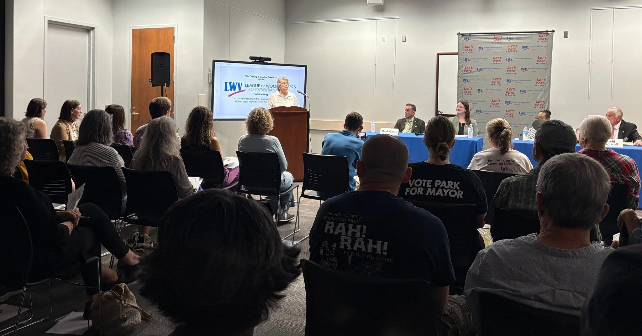 A panel of speakers at a community meeting, with an audience seated and listening. The speaker is at a podium, with a screen displaying the League of Women Voters logo behind her. The room has a gray wall with a large LWV banner.