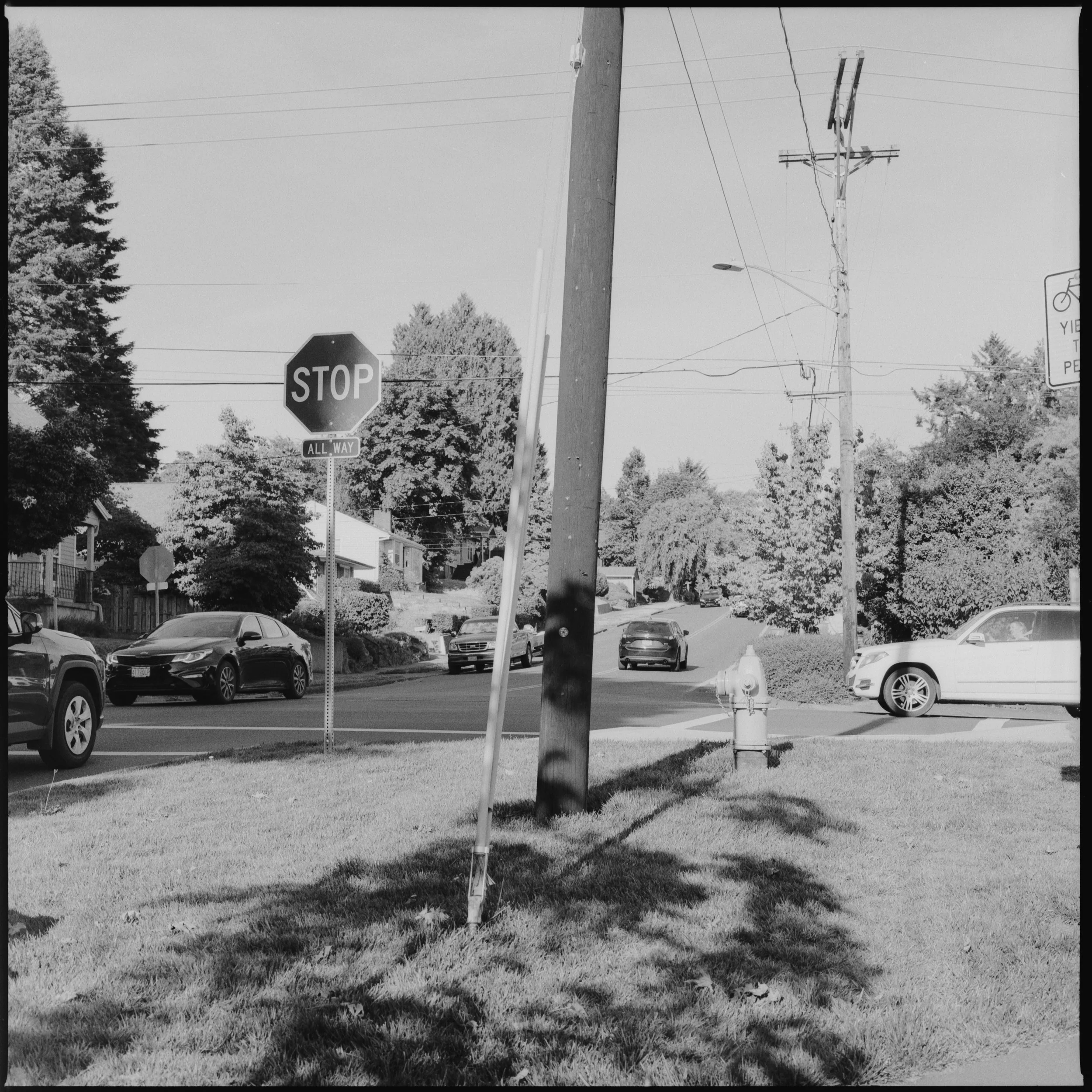 A black and white photo of a suburban street intersection with a stop sign, several parked and moving cars, utility poles, trees, and a fire hydrant.