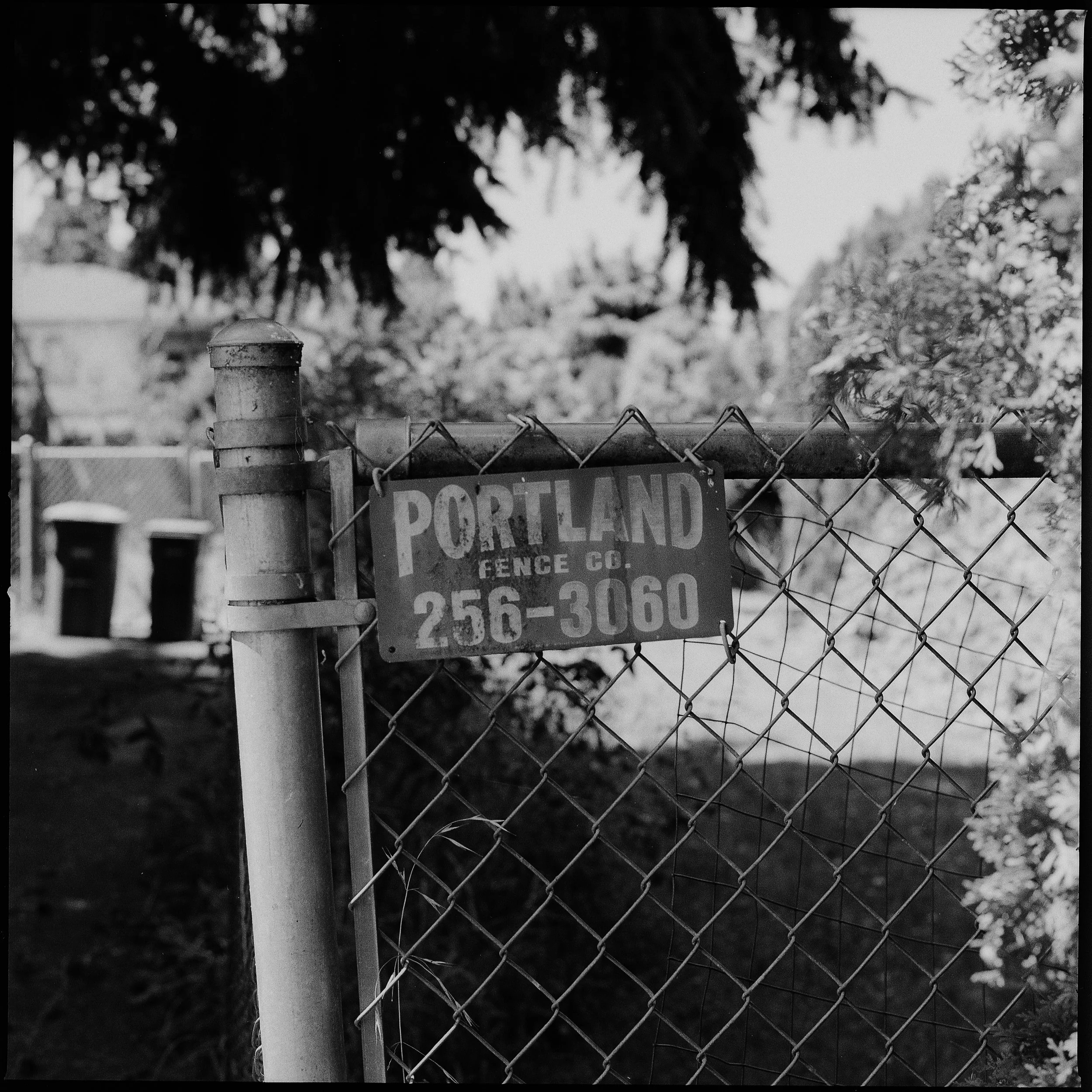 A black-and-white photograph of a chain-link fence with a Portland Fence Co. sign attached to it, displaying a phone number. Trees and some buildings are visible in the background.