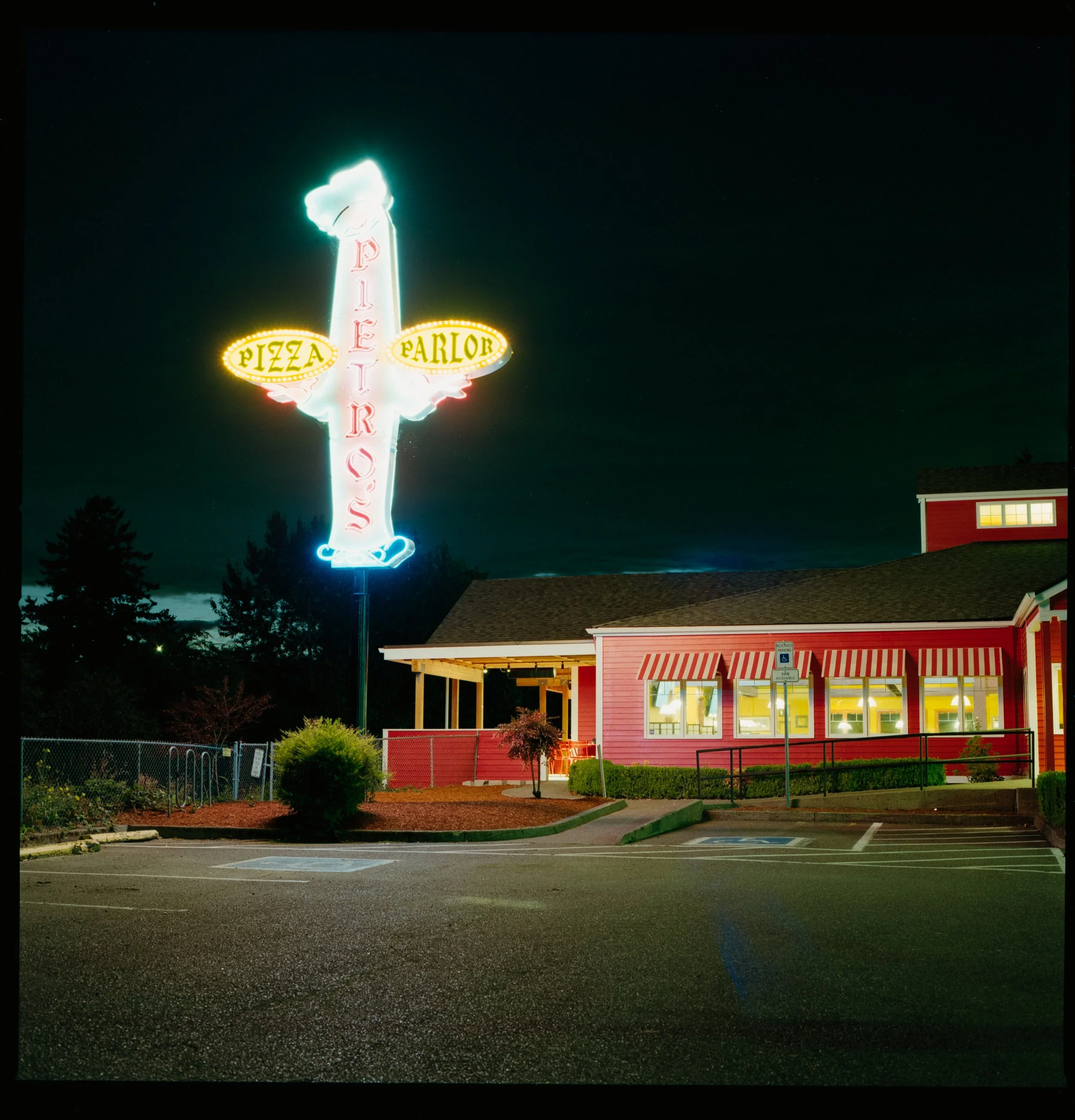 Nighttime exterior view of a pizza restaurant with a large neon sign featuring a chef's hat and the words "Pizza Parlor" with "Pizza" and "Parlor" on oval signs, and the building is painted red with striped awnings and lit windows.