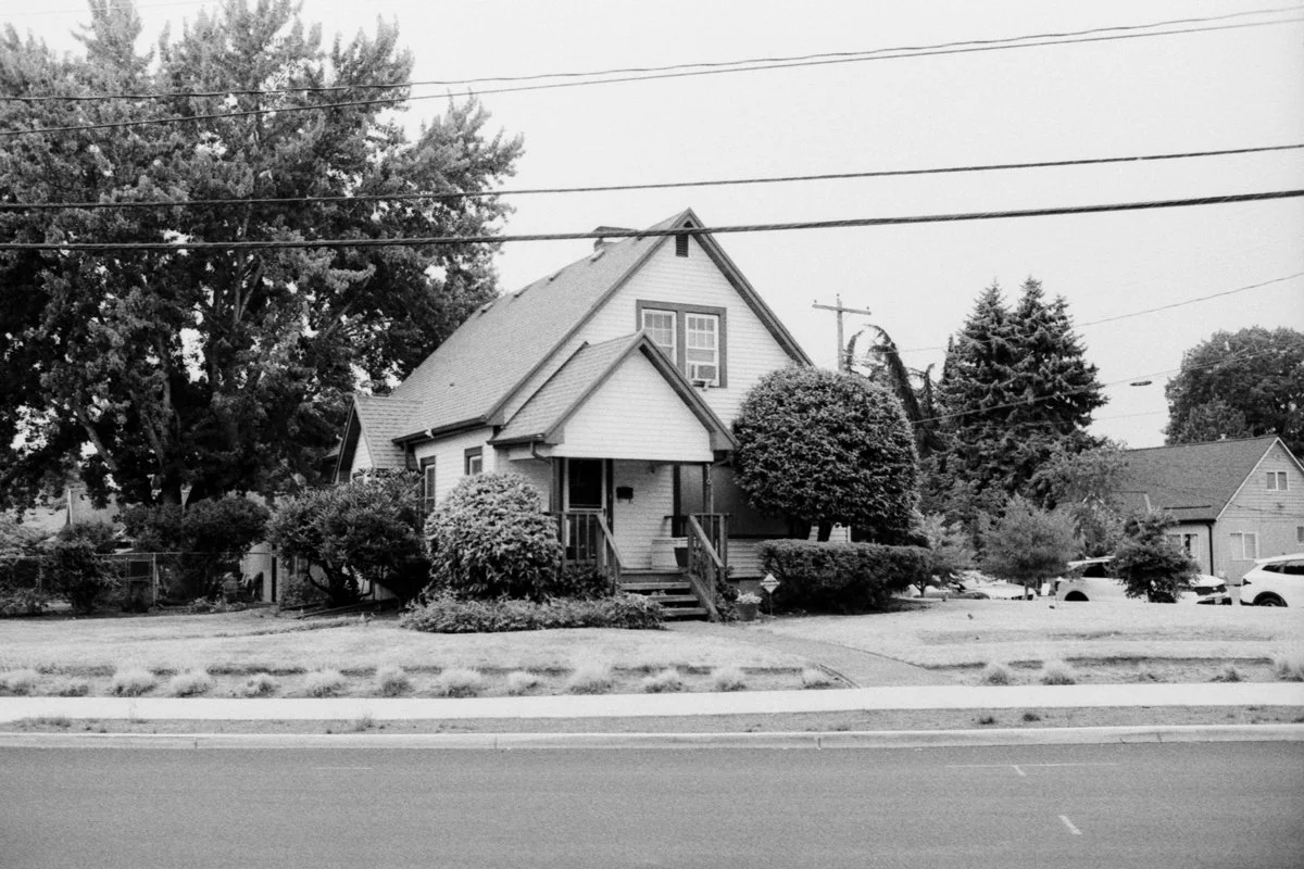 A black and white photo of a house with a gabled roof, front porch, and steps, surrounded by bushes and trees, with parked cars and other houses nearby.