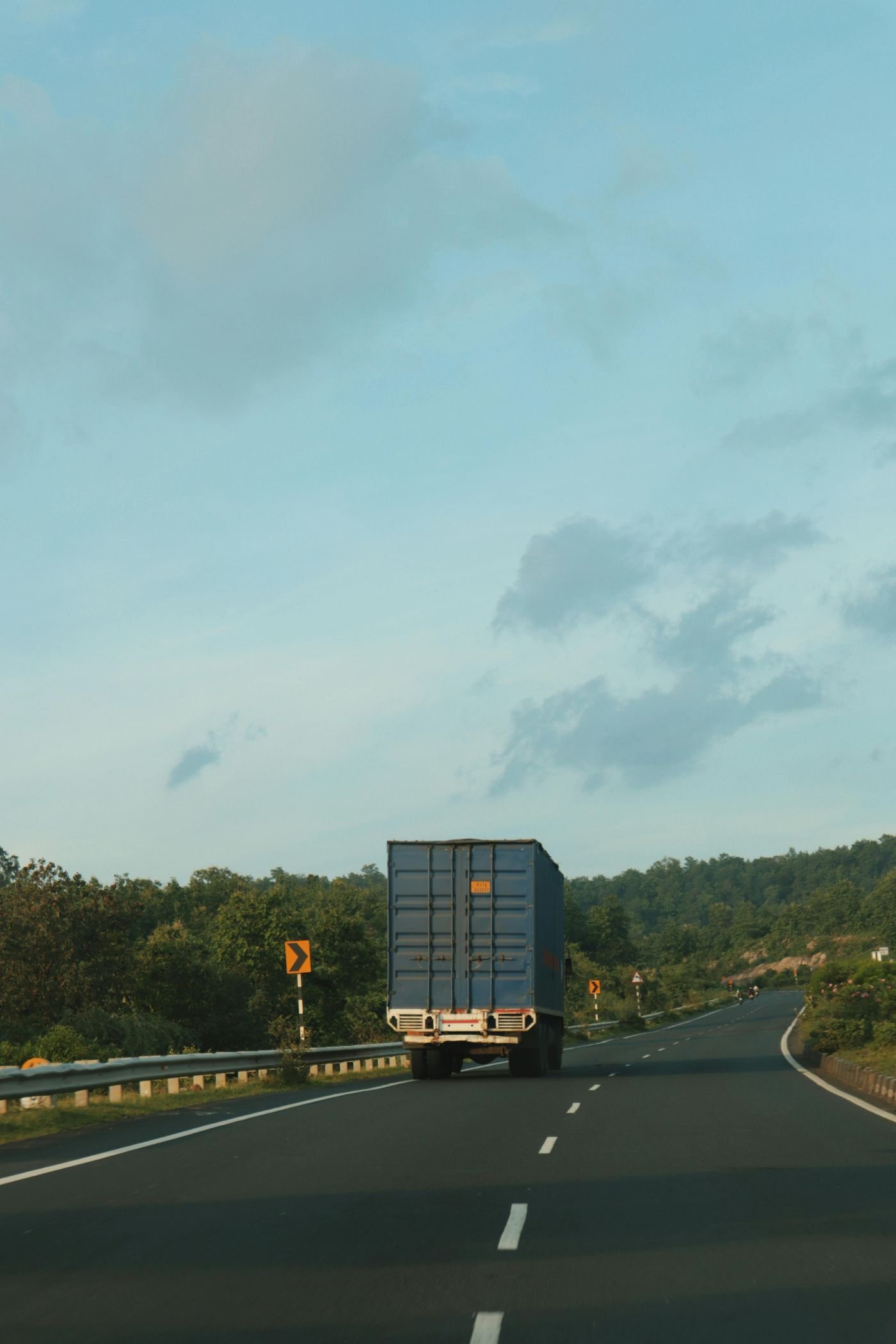 A blue container truck driving along a winding highway, surrounded by greenery and a clear sky with scattered clouds.