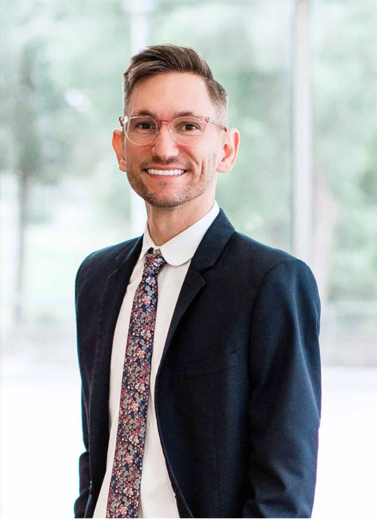 A smiling man with glasses, wearing a navy suit, white shirt, and a colorful floral tie, standing in a modern office with large windows and blurred outdoor scenery.