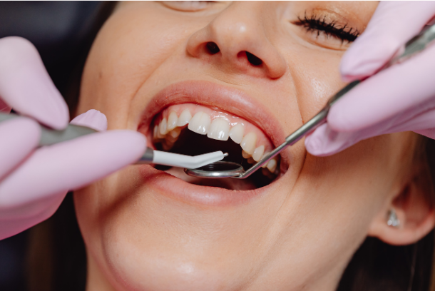 A woman receiving dental treatment with a dental mirror and suction tool in her mouth, smiling with her eyes closed.